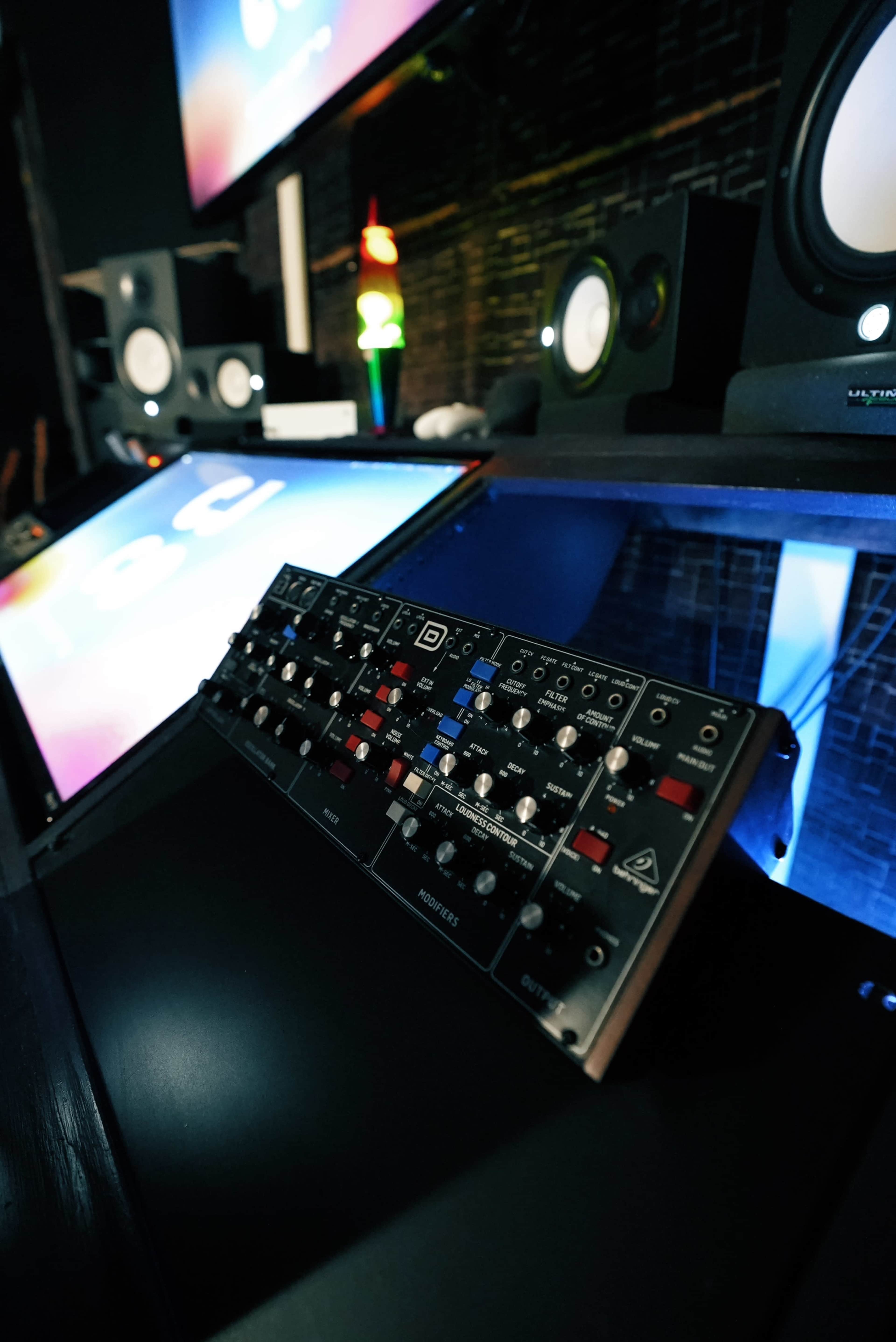 The image shows a close-up of an audio mixing console in a studio with monitors and colorful ambient lighting in the background.
