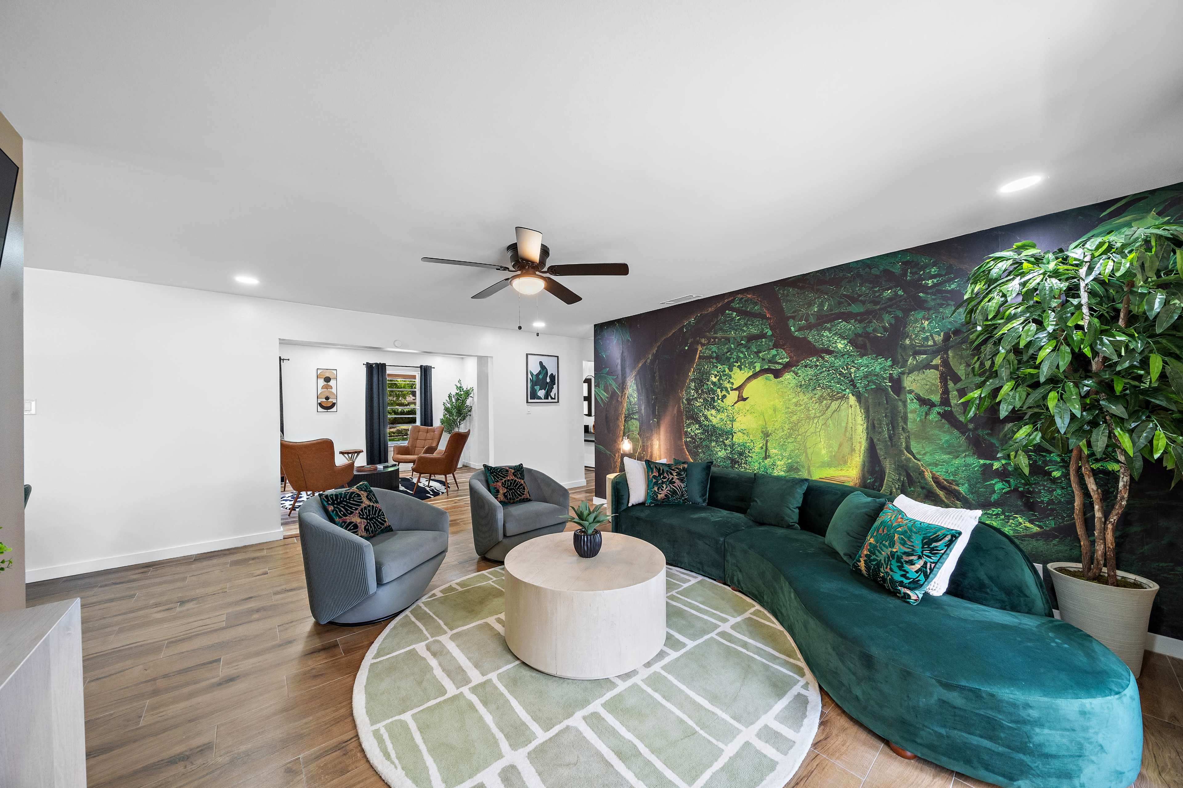 A modern living room featuring a curved green sofa, two armchairs, a round coffee table, and a mural of a forest on one wall, with a potted plant in the corner.