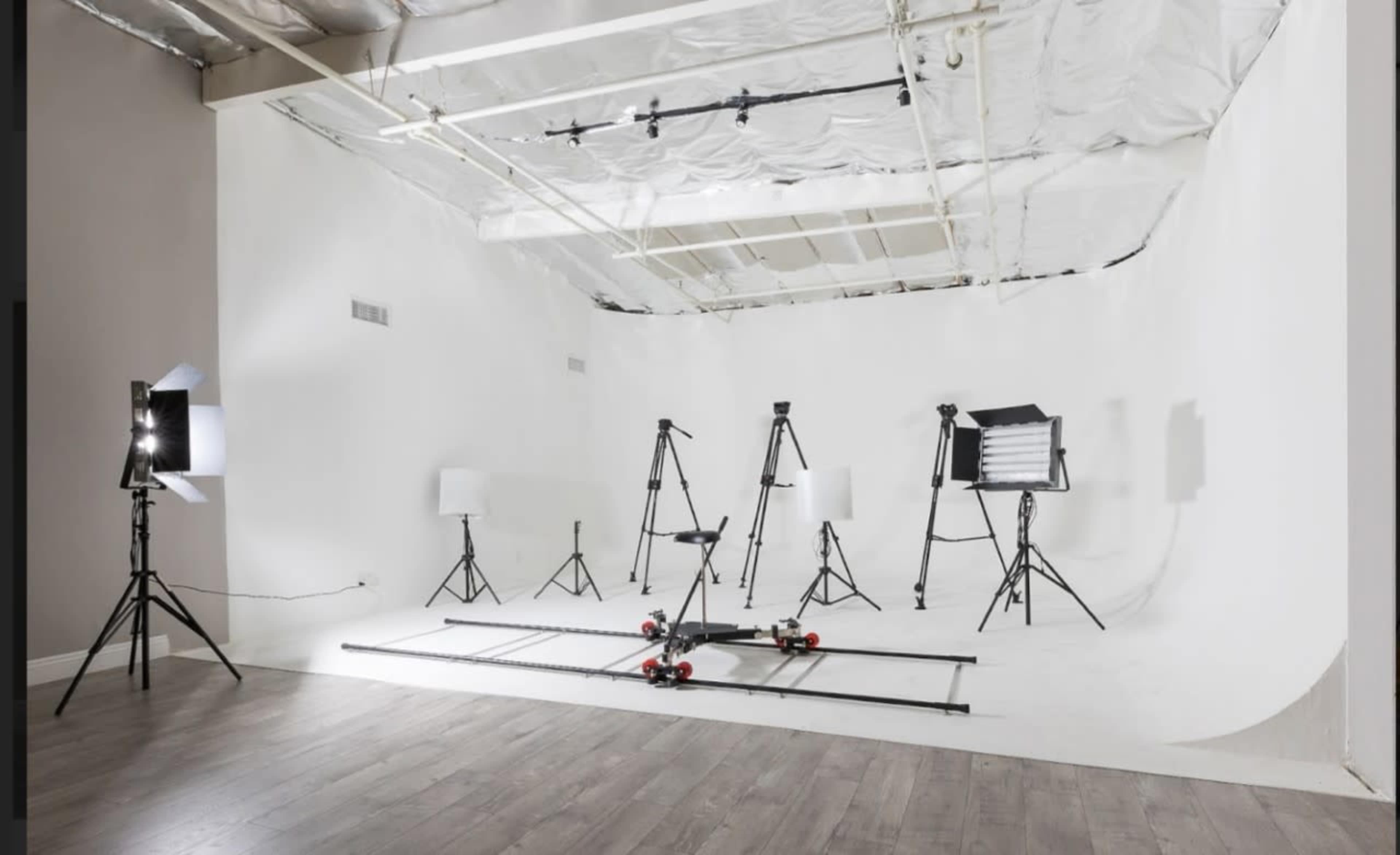 A minimalist studio setup with various photography equipment, including tripods, lighting fixtures, and a track for camera movement, arranged against a white backdrop.