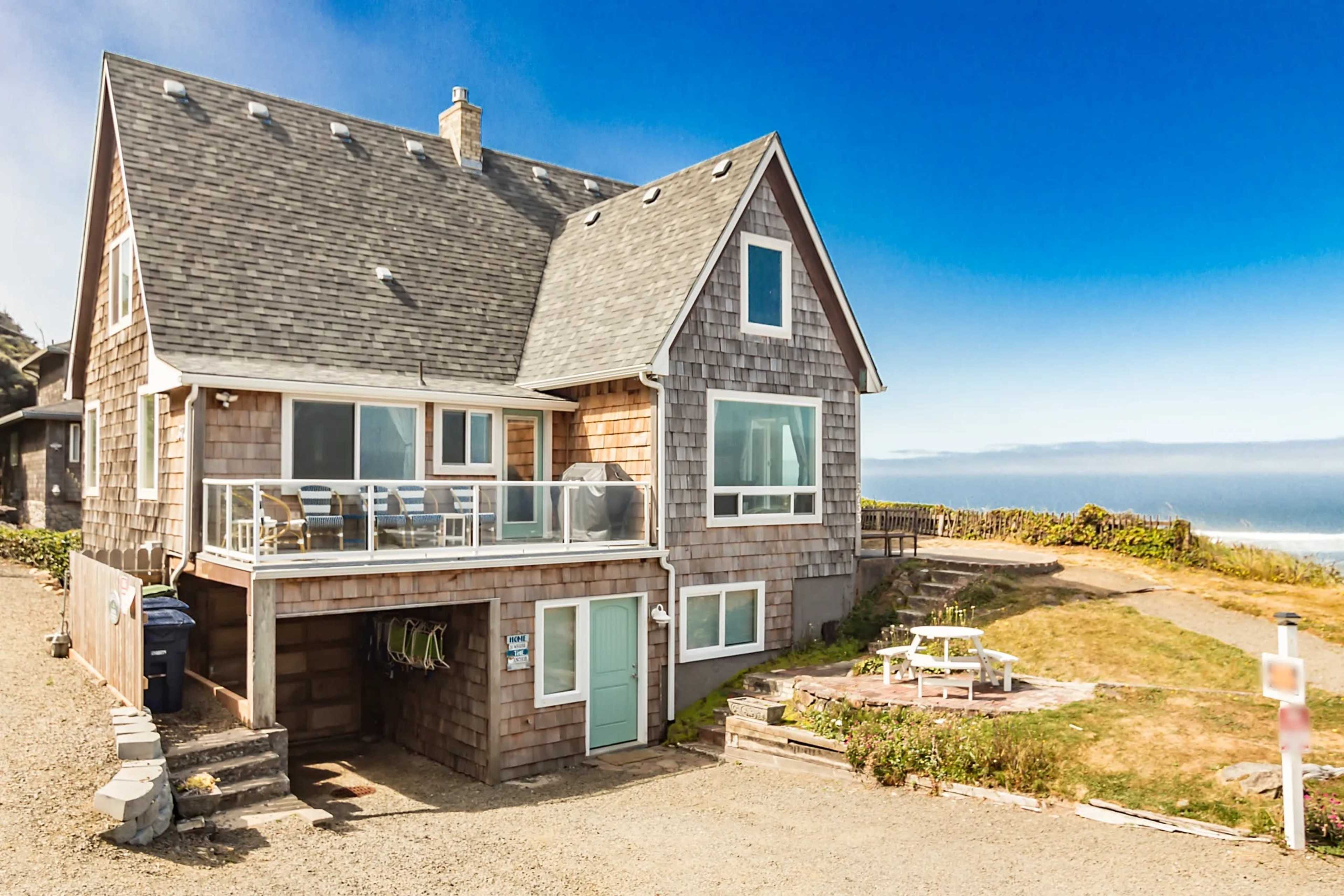 The image shows a two-story seaside house with wooden shingles and a balcony overlooking a grassy area and the ocean.