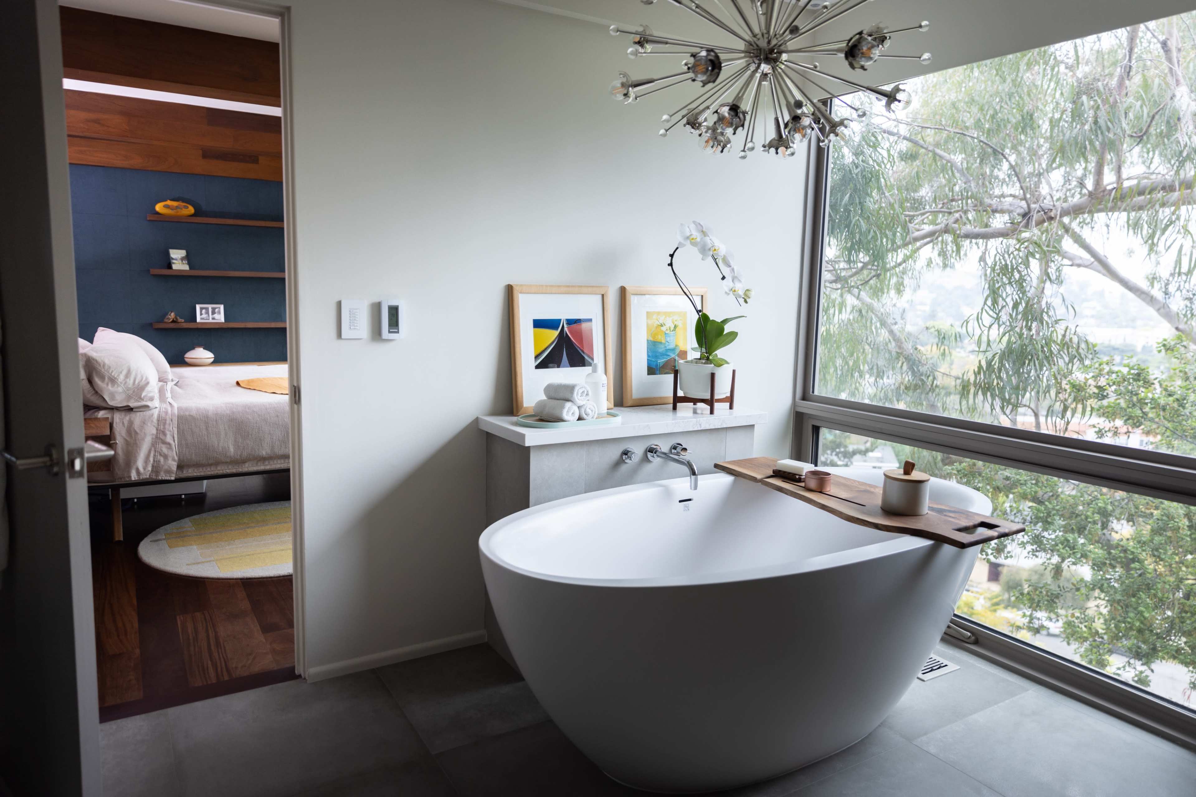 The image shows a modern bathroom featuring a sleek, freestanding bathtub placed near a large window with views of greenery.