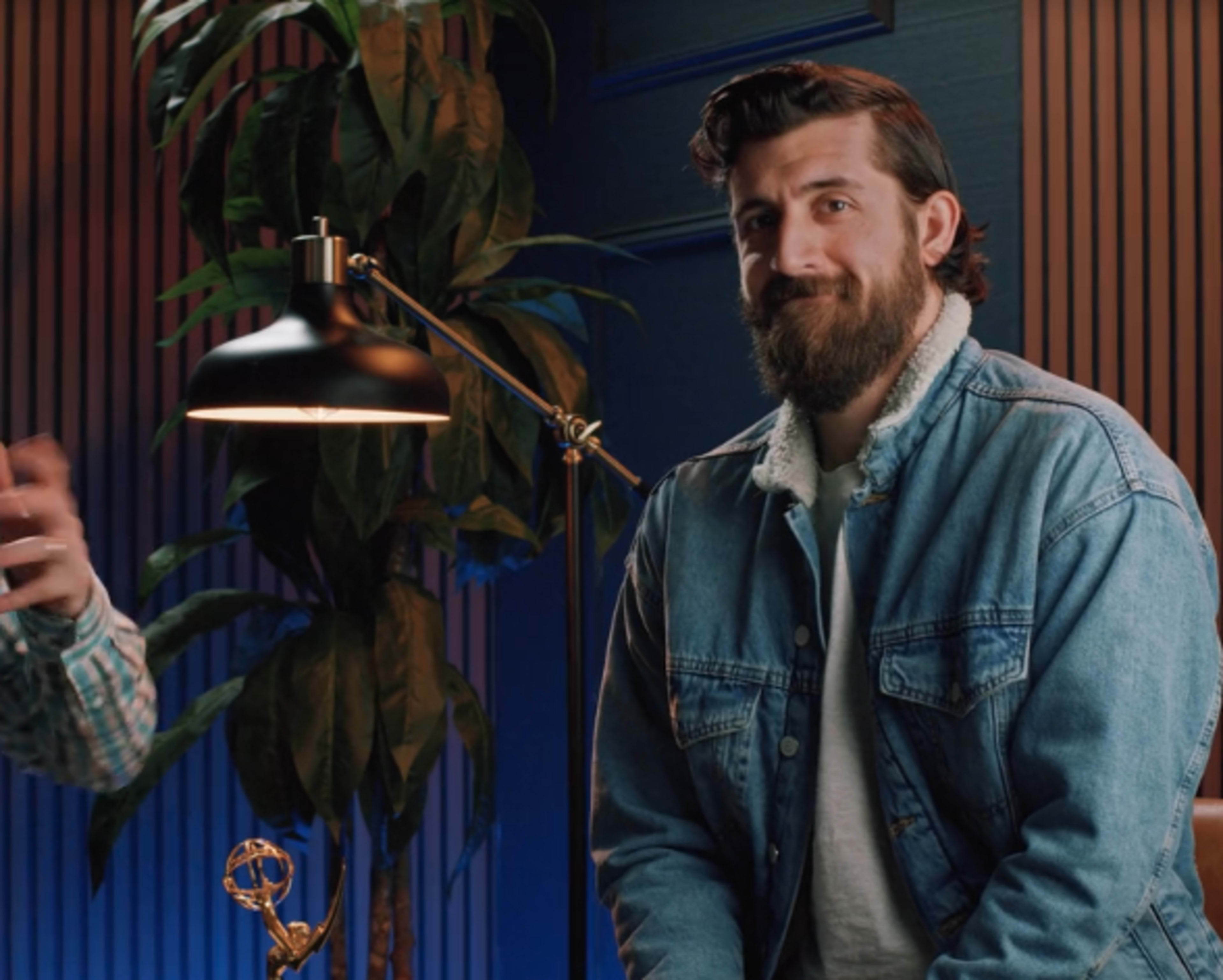A man with a beard is sitting next to a lamp and a potted plant in a room decorated with blue walls and wooden paneling.