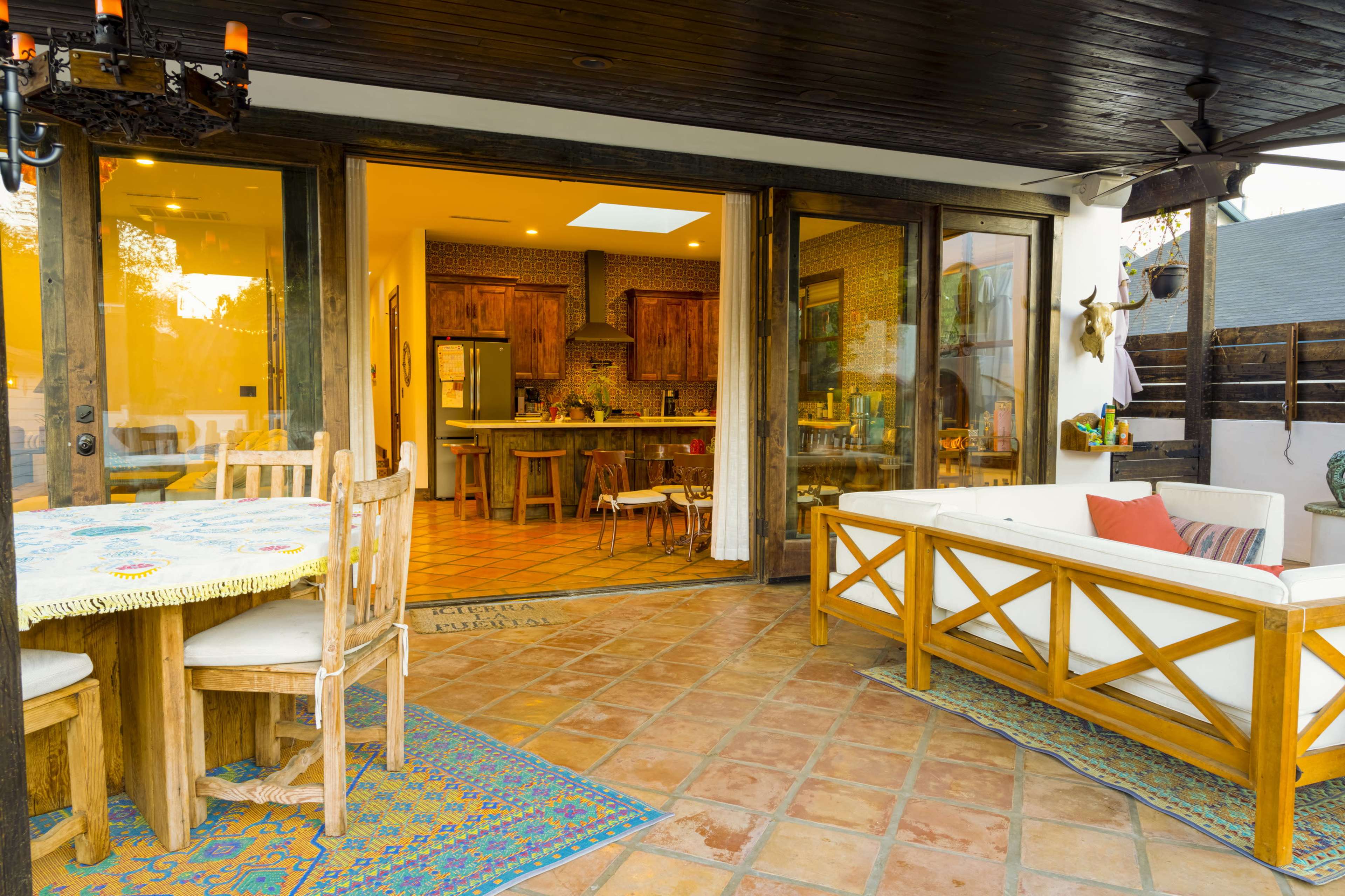 A cozy outdoor dining area features a wooden table and chairs, with a view of a kitchen through large glass doors, all set on a tiled patio.