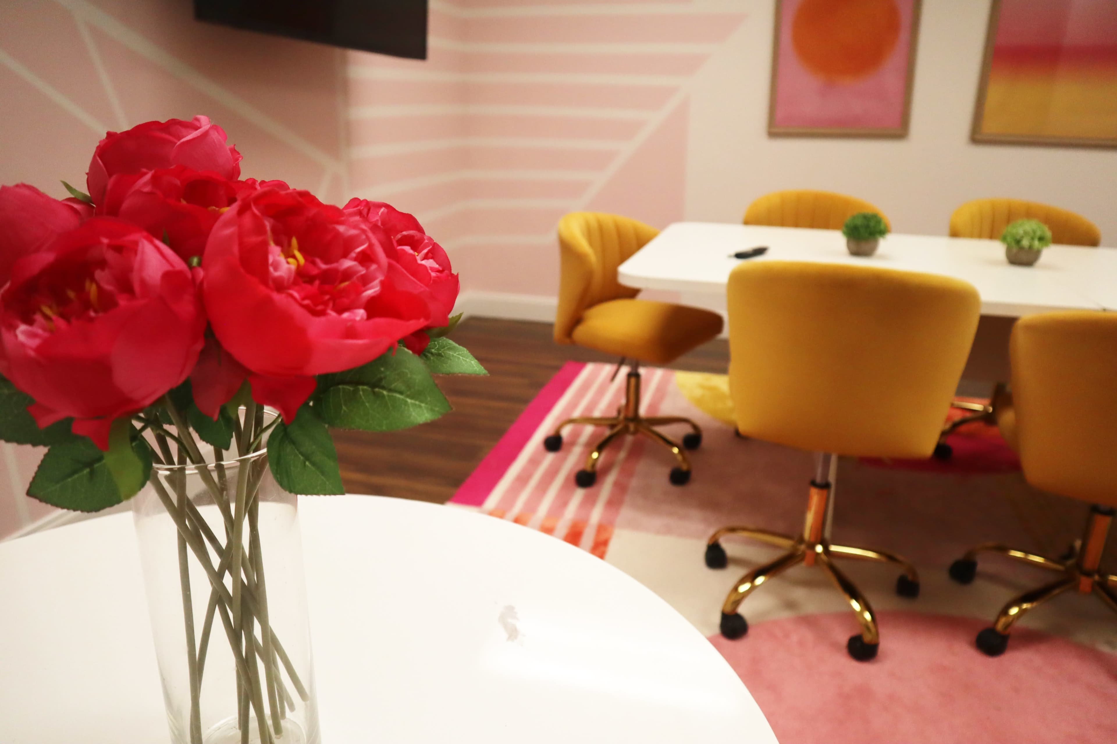 A conference room featuring a decorated white table, yellow chairs, pink flower arrangements, and colorful artwork on the walls.