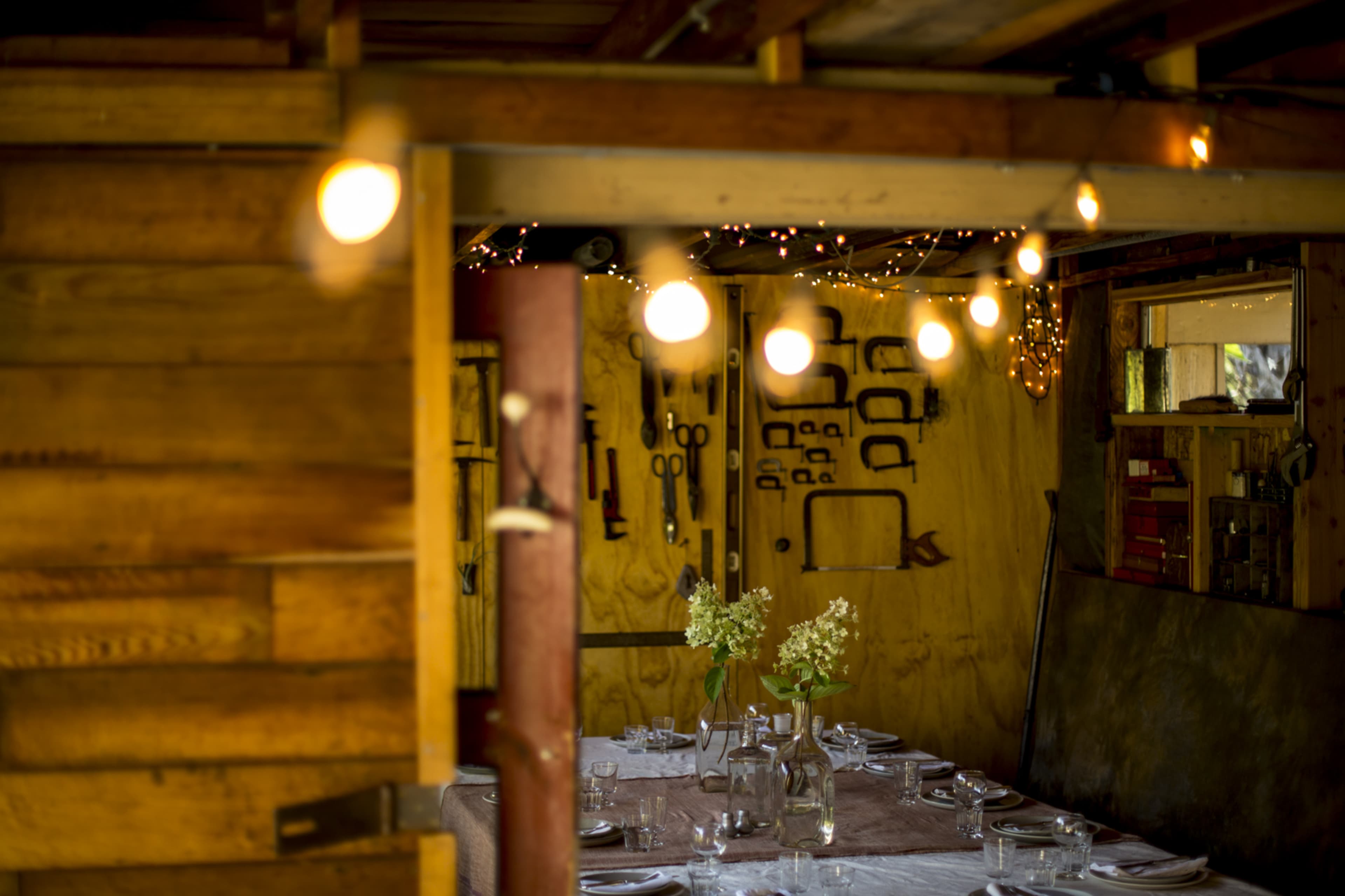 A rustic wooden interior features a dining table set with glassware and flowers, illuminated by string lights overhead.