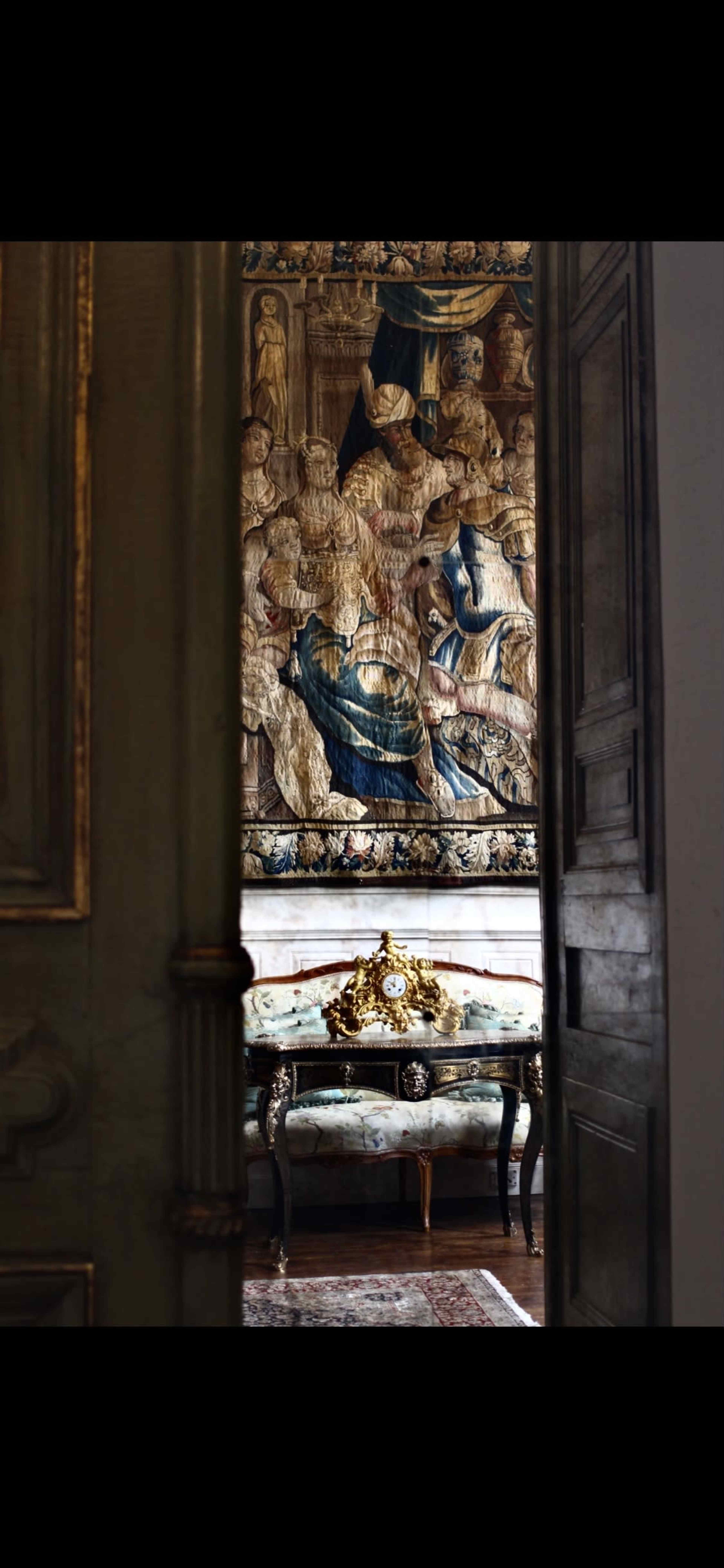 A view through an open door reveals an ornate room featuring a decorated wall tapestry and a vintage sofa with a clock on a side table.