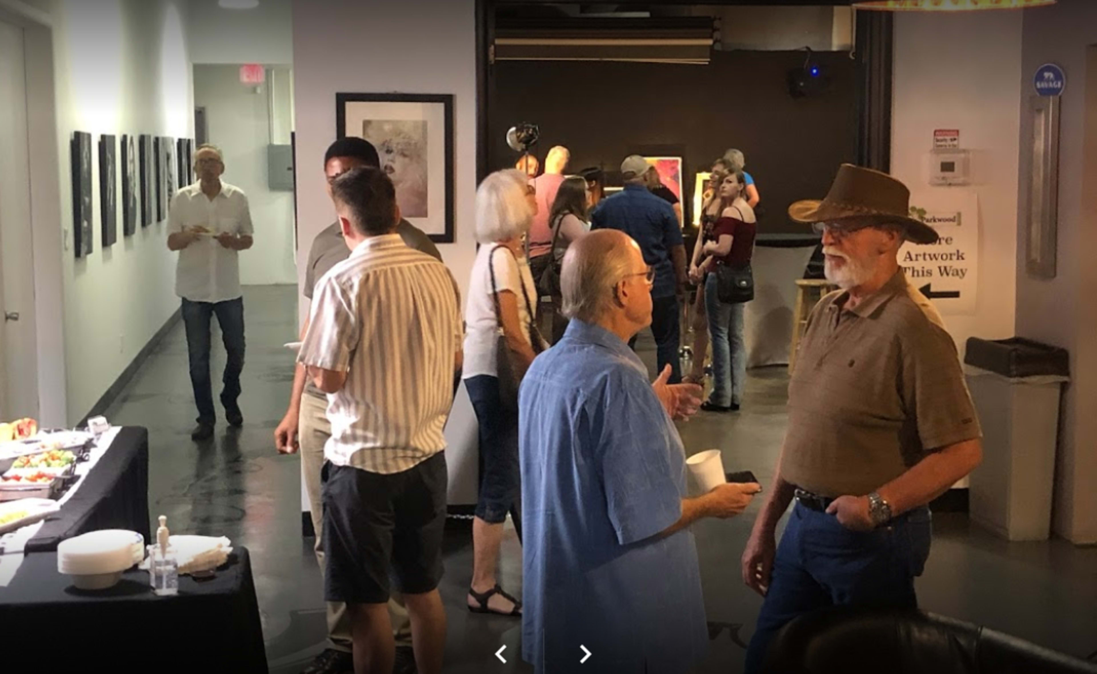 A group of people mingles at an art gallery event, with a buffet table in the foreground and artwork displayed on the walls.