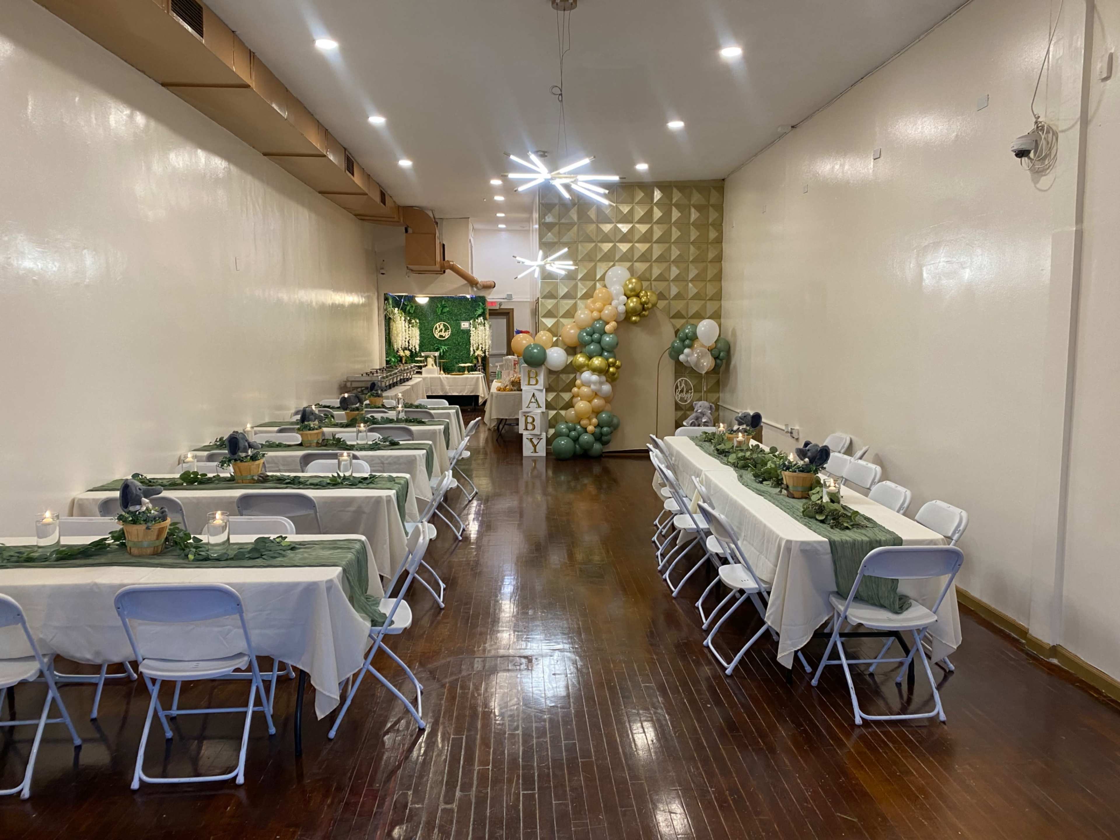 The photo shows a decorated event space with tables set up neatly on either side, a balloon display in the back, and a food serving area along one wall.