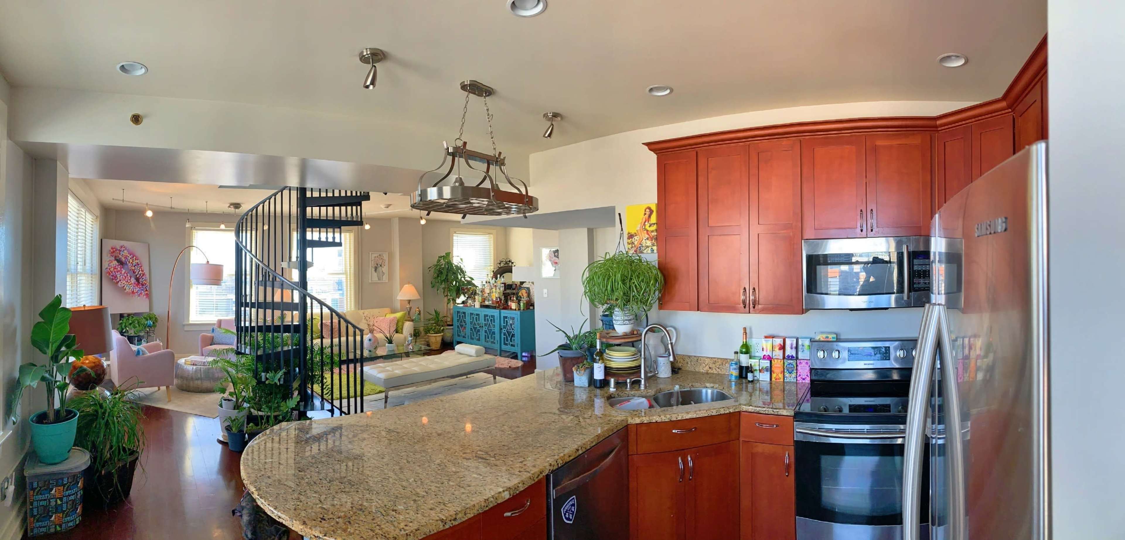 A modern open-concept kitchen with granite countertops and stainless steel appliances, seamlessly connected to a living area featuring a spiral staircase and various plants.