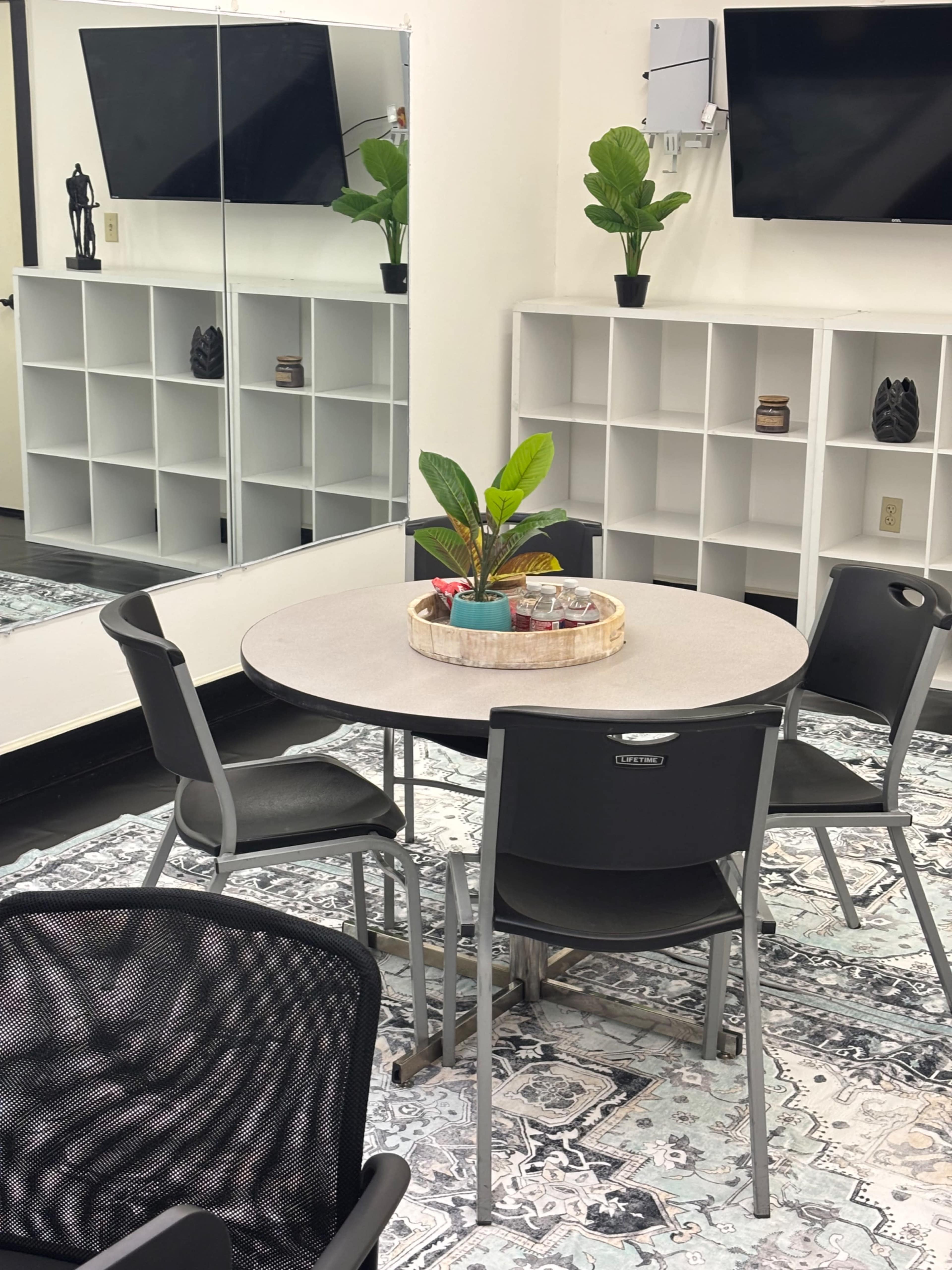A small meeting room features a round table with a plant centerpiece, surrounded by black chairs, with shelves and two TVs visible in the background.