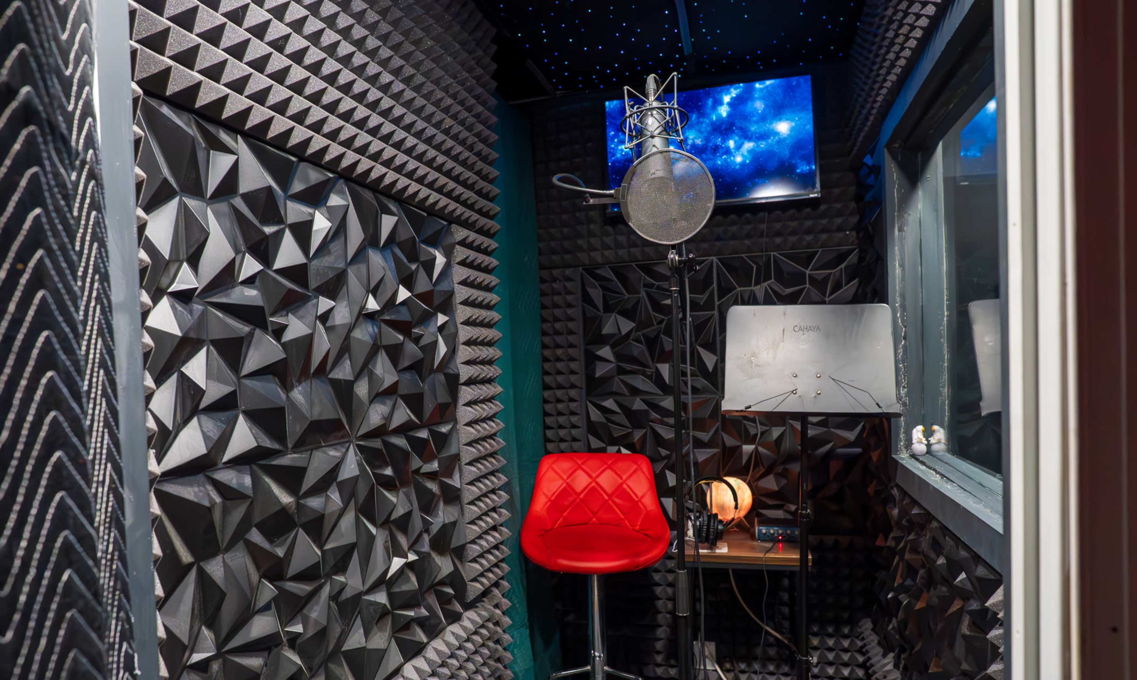 The image shows a soundproof recording booth with textured wall panels, a microphone on a stand, a red chair, and a small table with equipment.