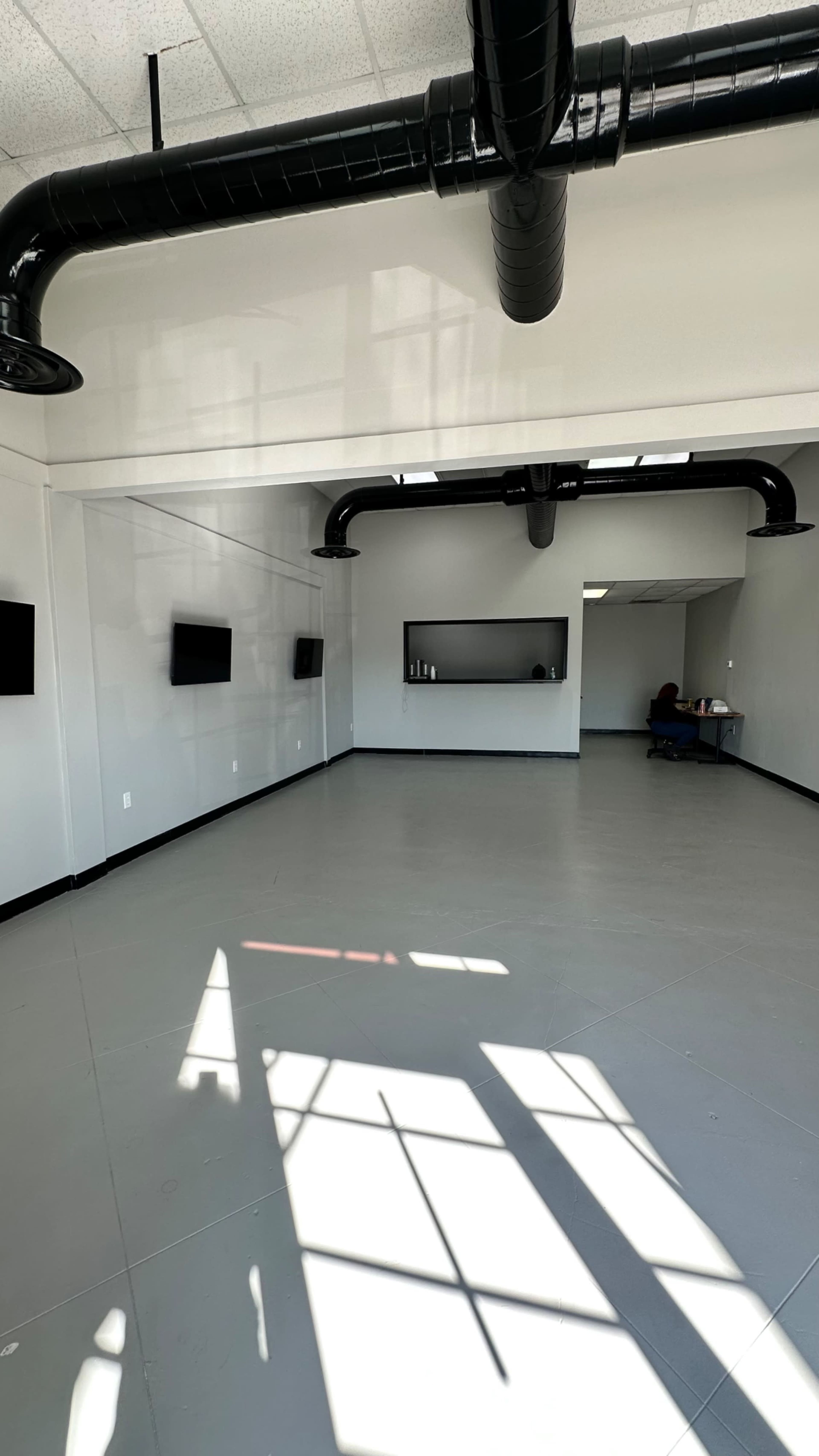The image shows a spacious, empty room with gray flooring, high ceilings, and black ventilation pipes, featuring several wall-mounted screens and a desk in the corner.