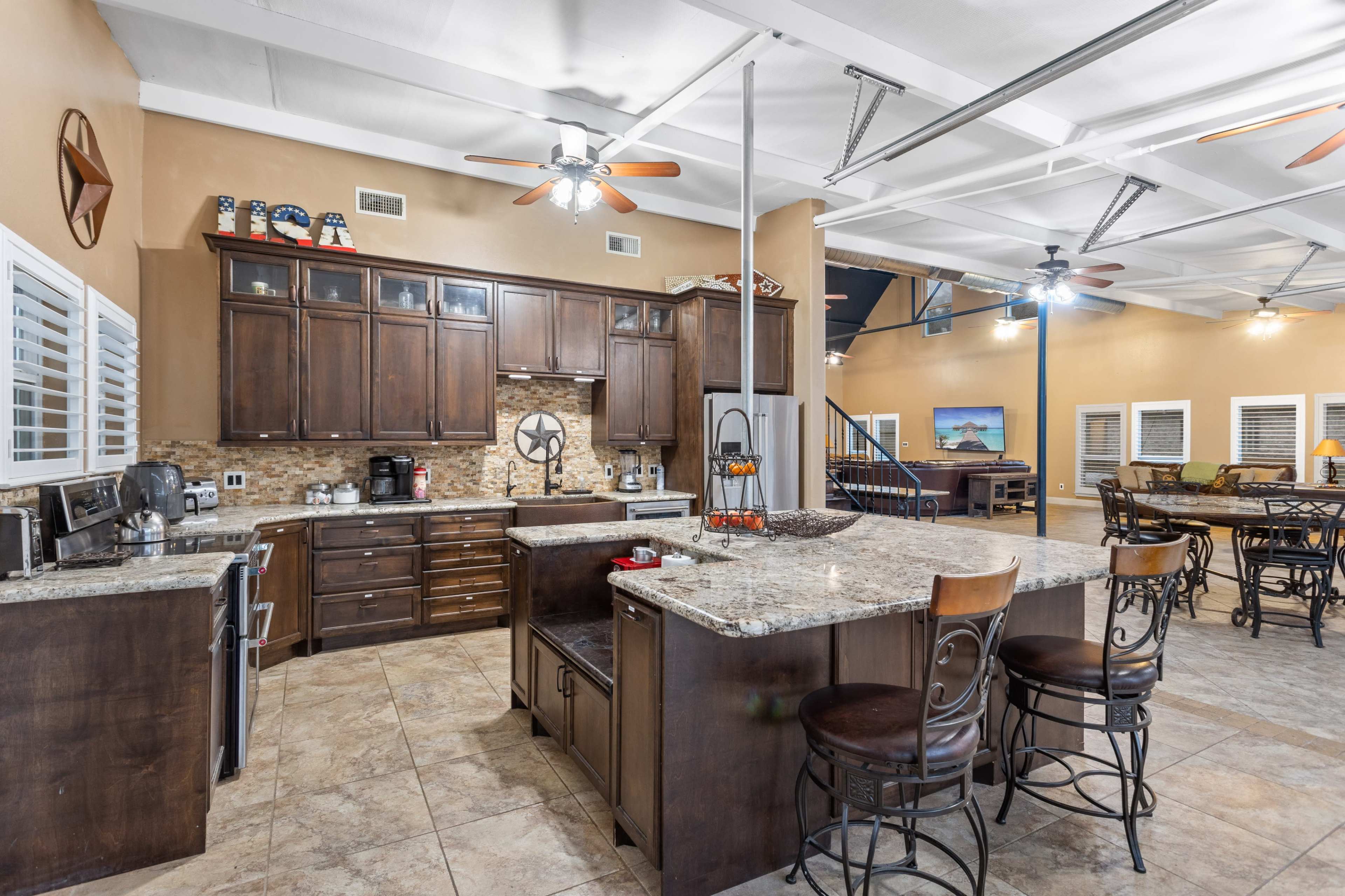 A spacious kitchen with dark wooden cabinets, granite countertops, and an island with bar stools, adjacent to a large open living area.