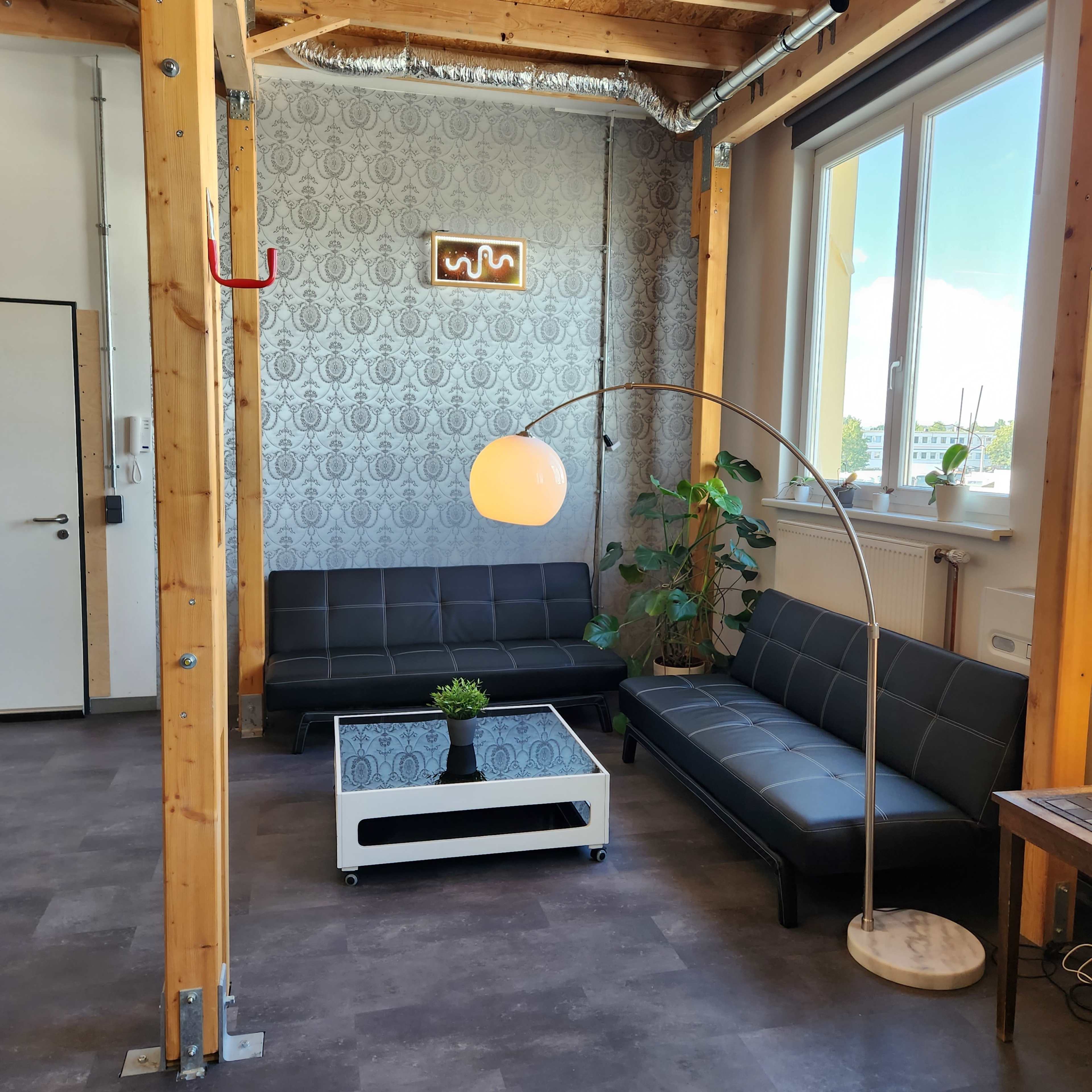 The image shows a modern lounge area with two black sofas, a round floor lamp, a coffee table, and greenery, set against a patterned wall and large windows.