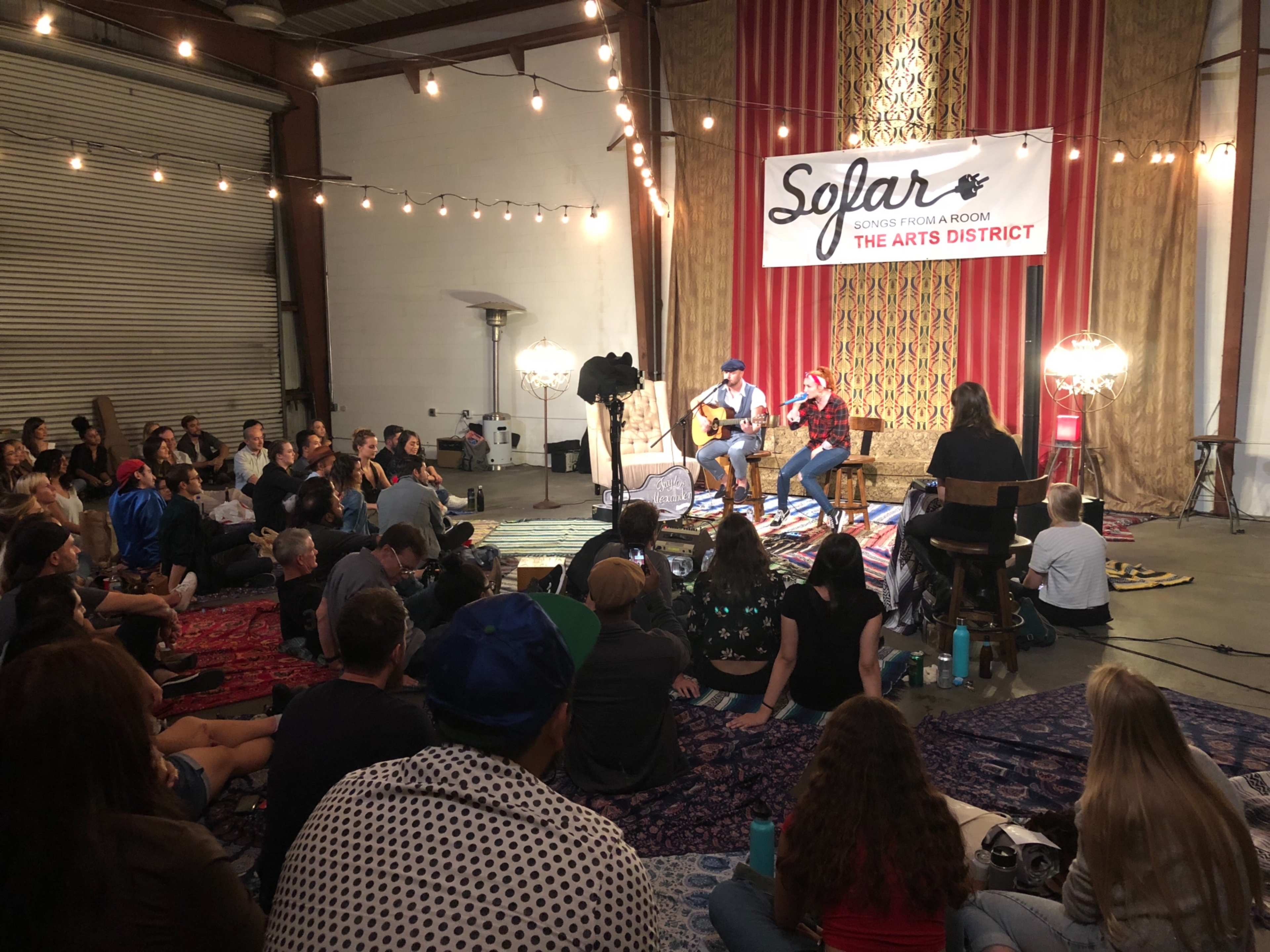 A crowd sits on the floor of a venue, watching two musicians perform on stage with acoustic guitars under warm lighting.
