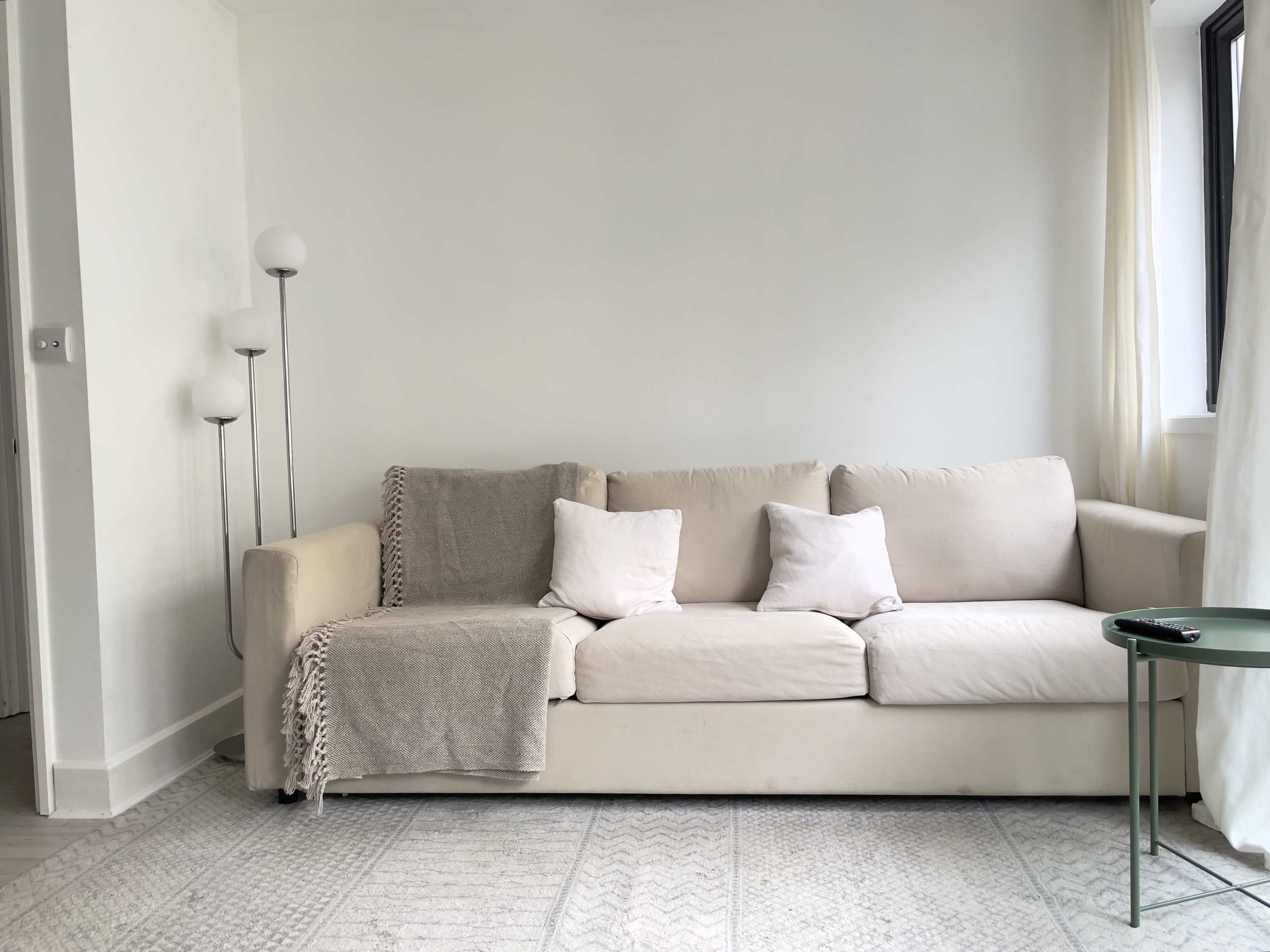 A light-colored sofa with a throw blanket and several pillows, positioned in a minimalistic room with a floor lamp and a small green side table.