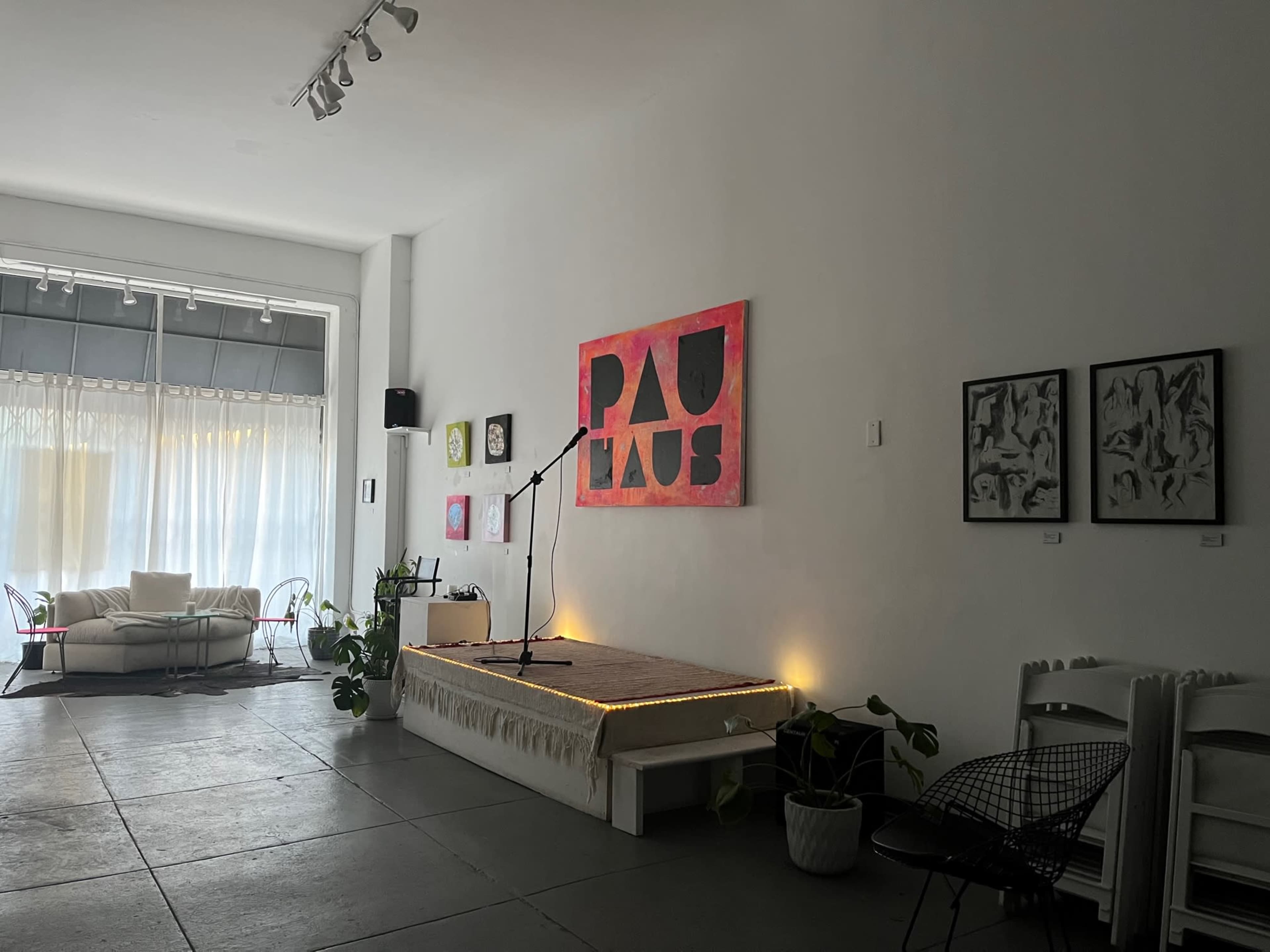 A minimalist performance space with a microphone on a small stage, surrounded by plants and artwork on the walls.
