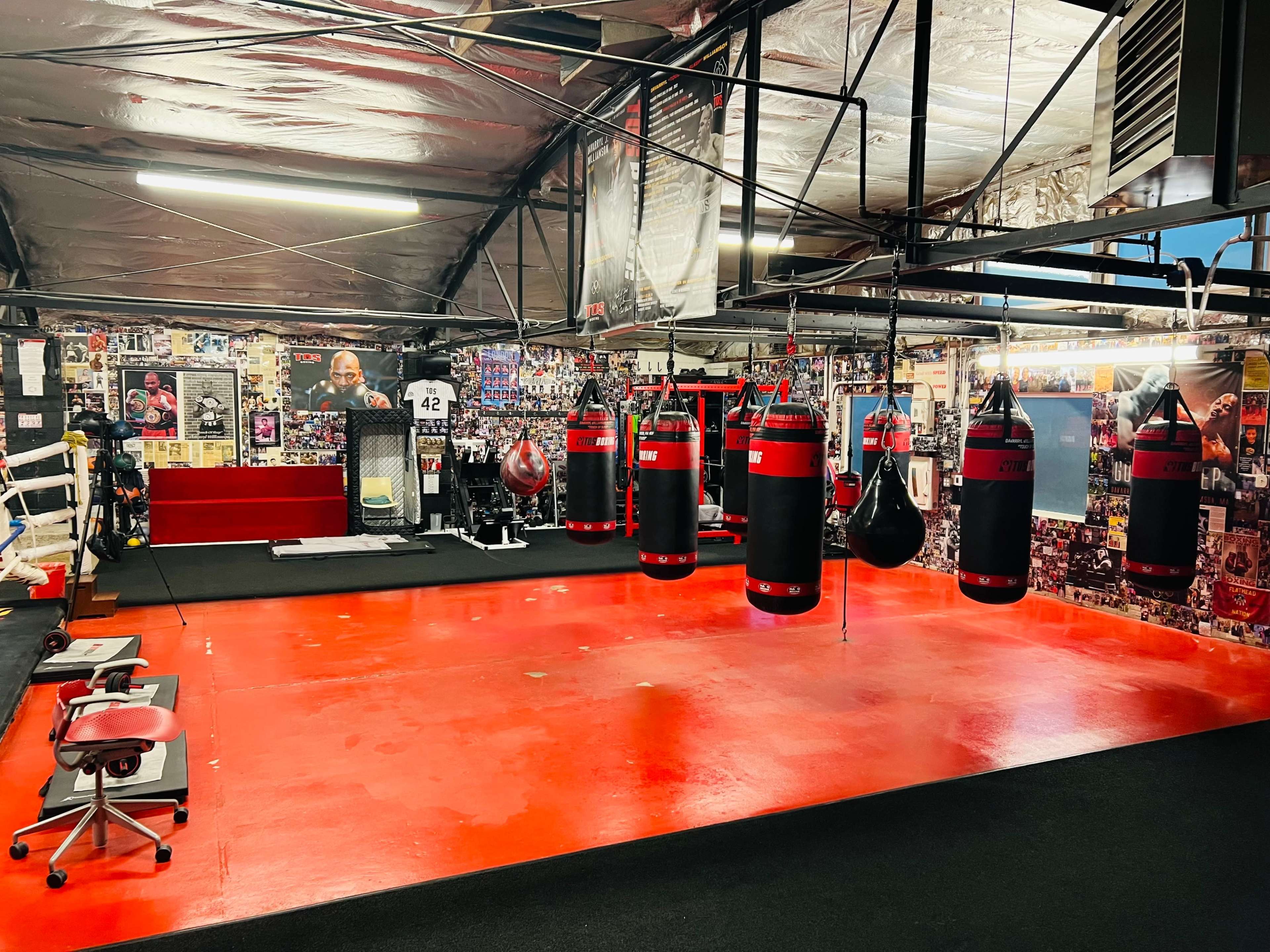 The image shows a boxing gym with red flooring, several hanging punching bags, and a collage of photographs on the walls.