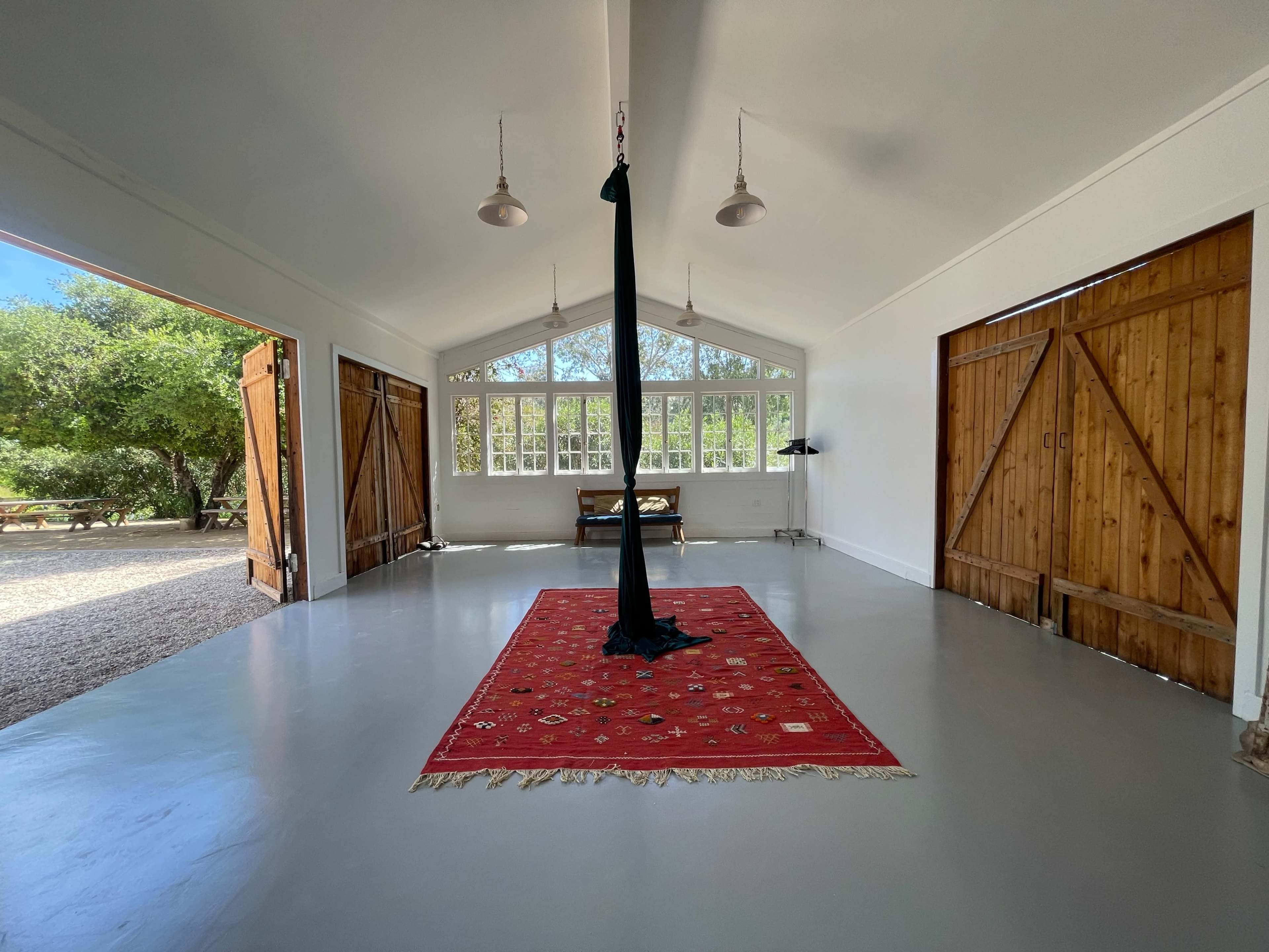 The interior of a spacious, well-lit room features a red patterned rug centered on a polished gray floor, with large windows and two wooden doors open to a view of greenery outside.