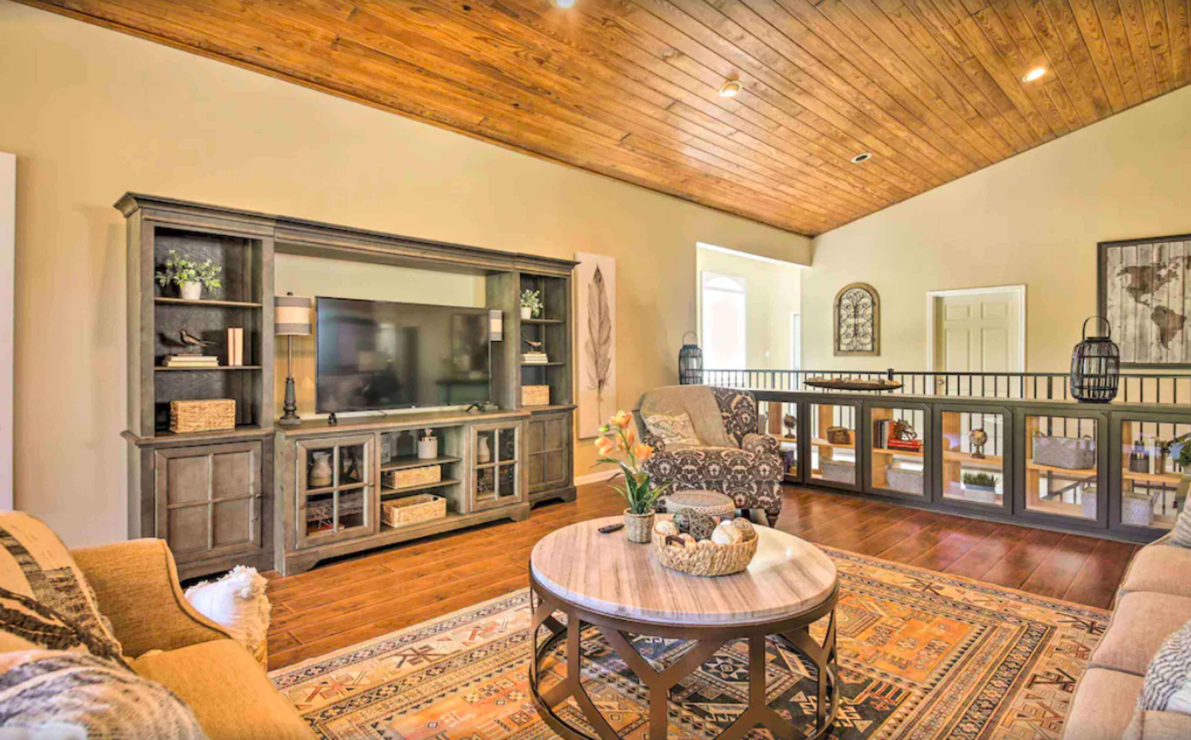 A living room with a wooden ceiling, a large TV unit, a round coffee table, and comfortable seating arranged around a patterned area rug.