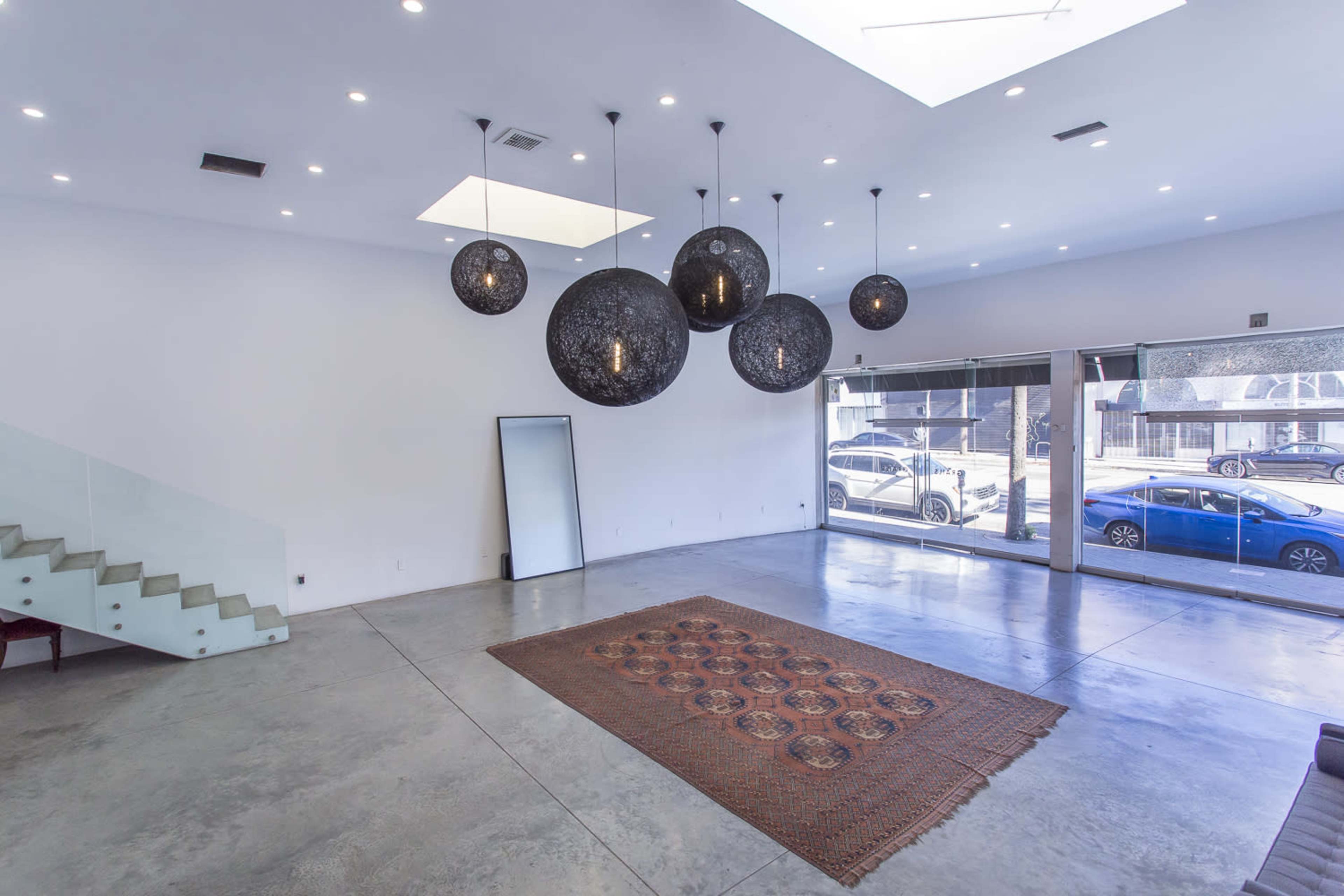 The image shows a spacious room with a minimalist design, featuring a large rug on the concrete floor, modern pendant lights, and windows that provide natural light.