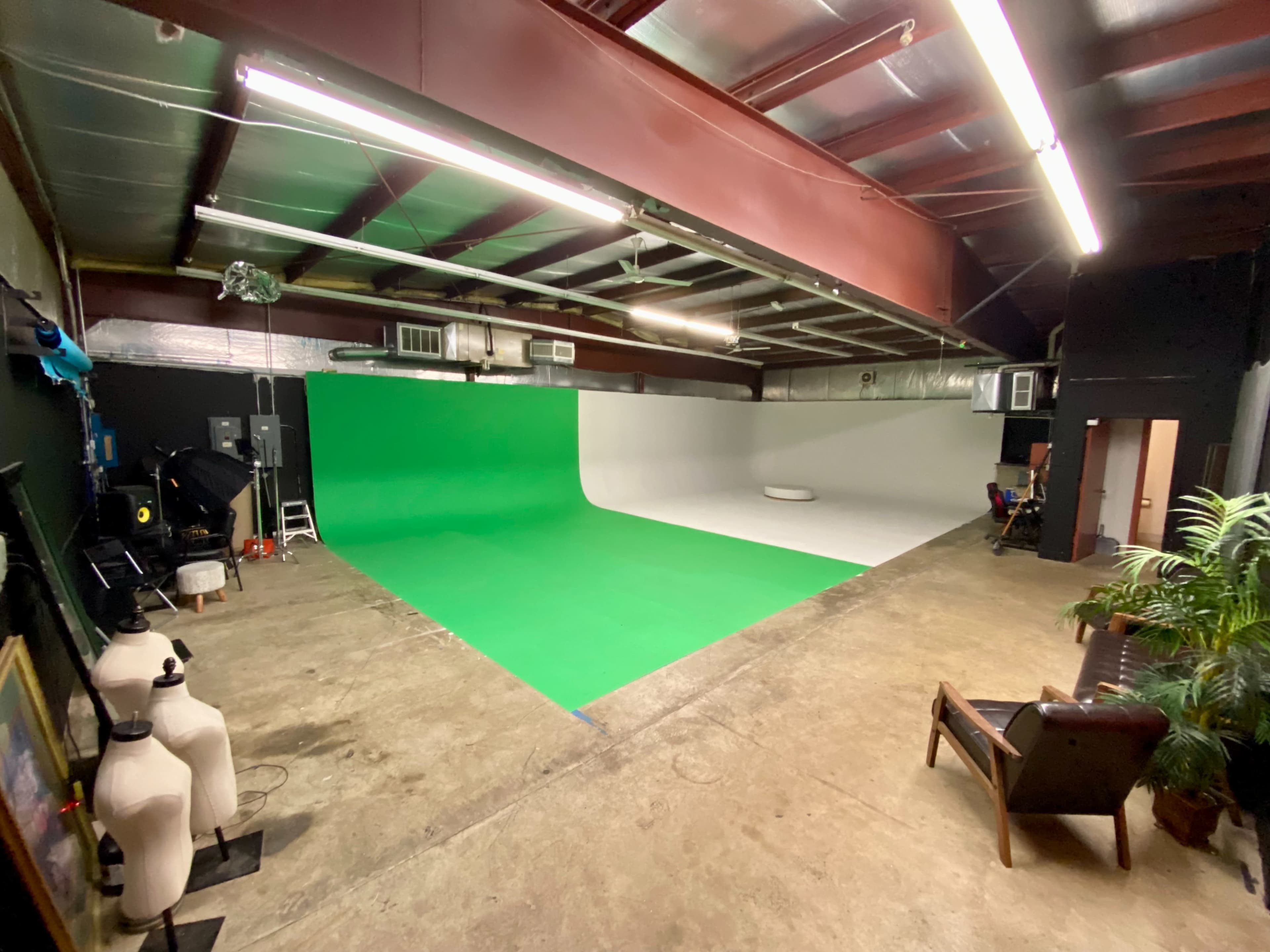 A spacious film studio features a green screen and a white backdrop, with equipment and furniture arranged along the walls.