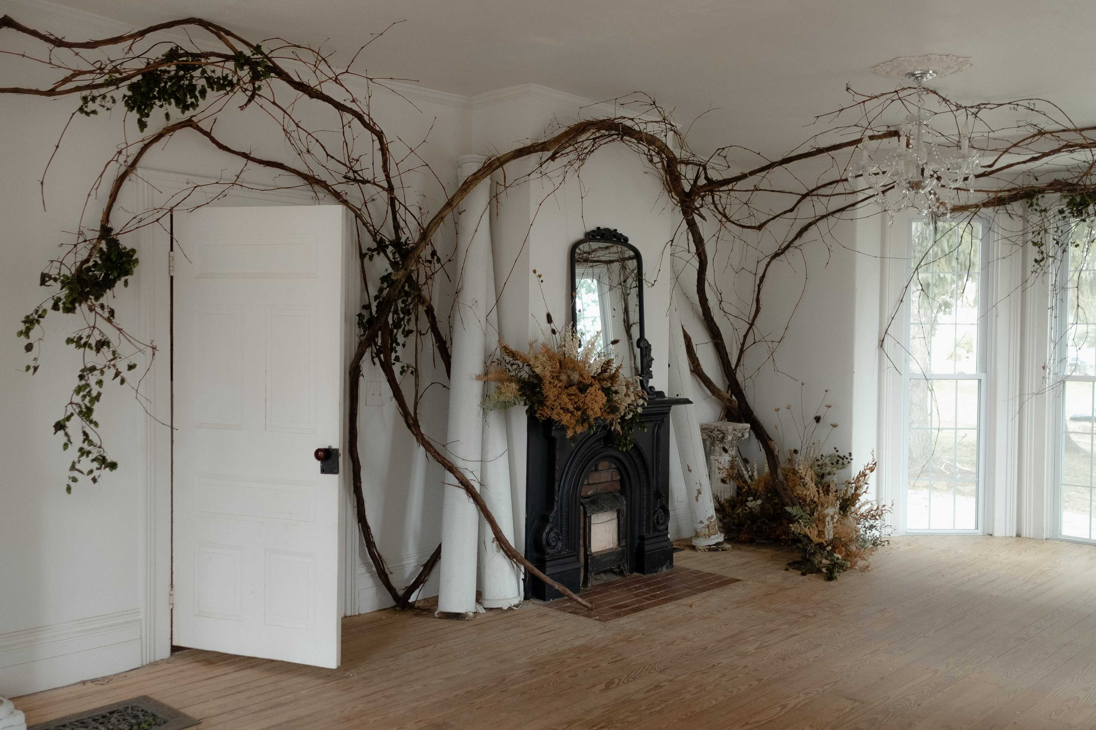 The interior features a fireplace surrounded by twisting branches and greenery, with a large mirror above it and light pouring in through the windows.