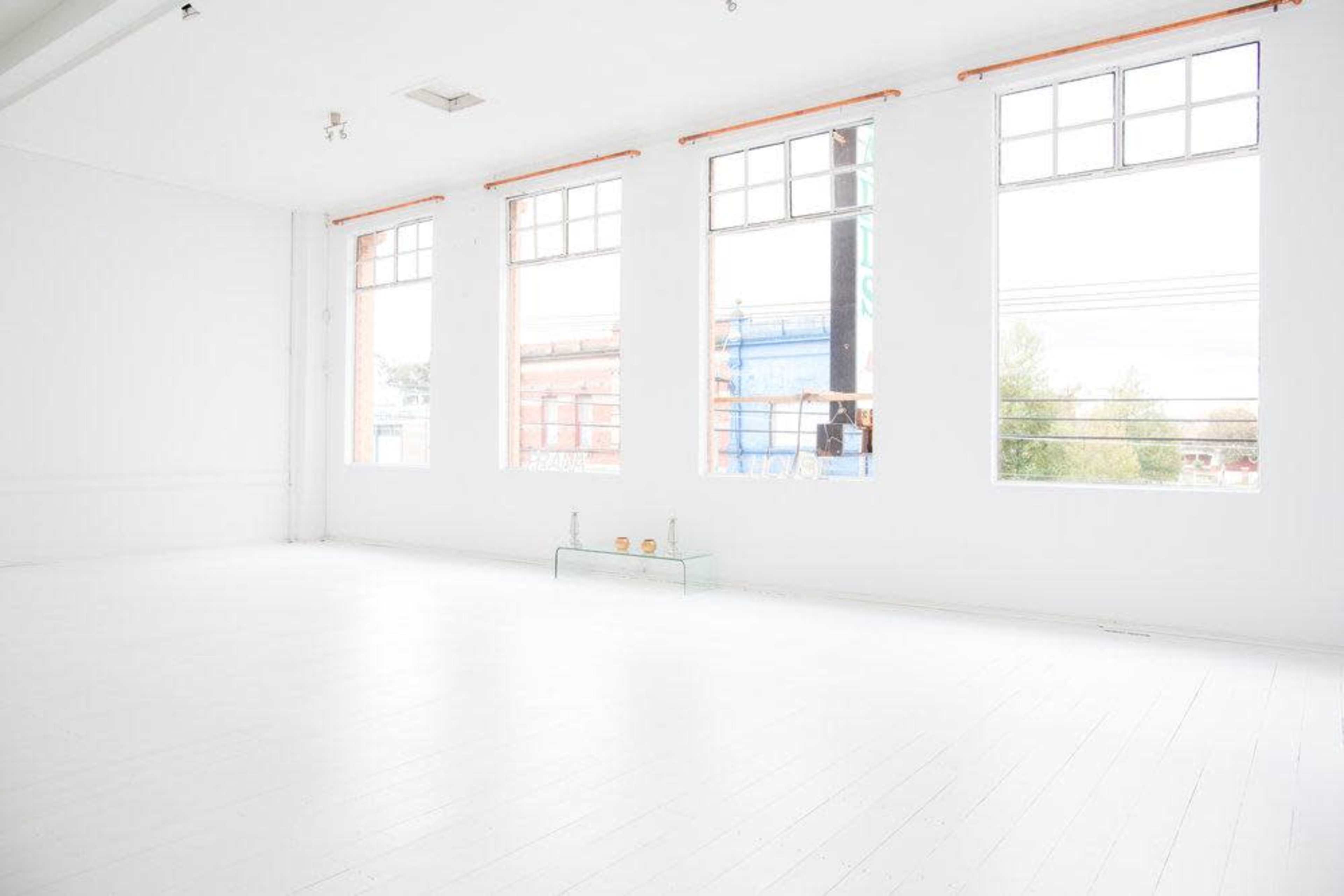 The image shows a bright, empty room with white walls and floors, featuring large windows that let in natural light.