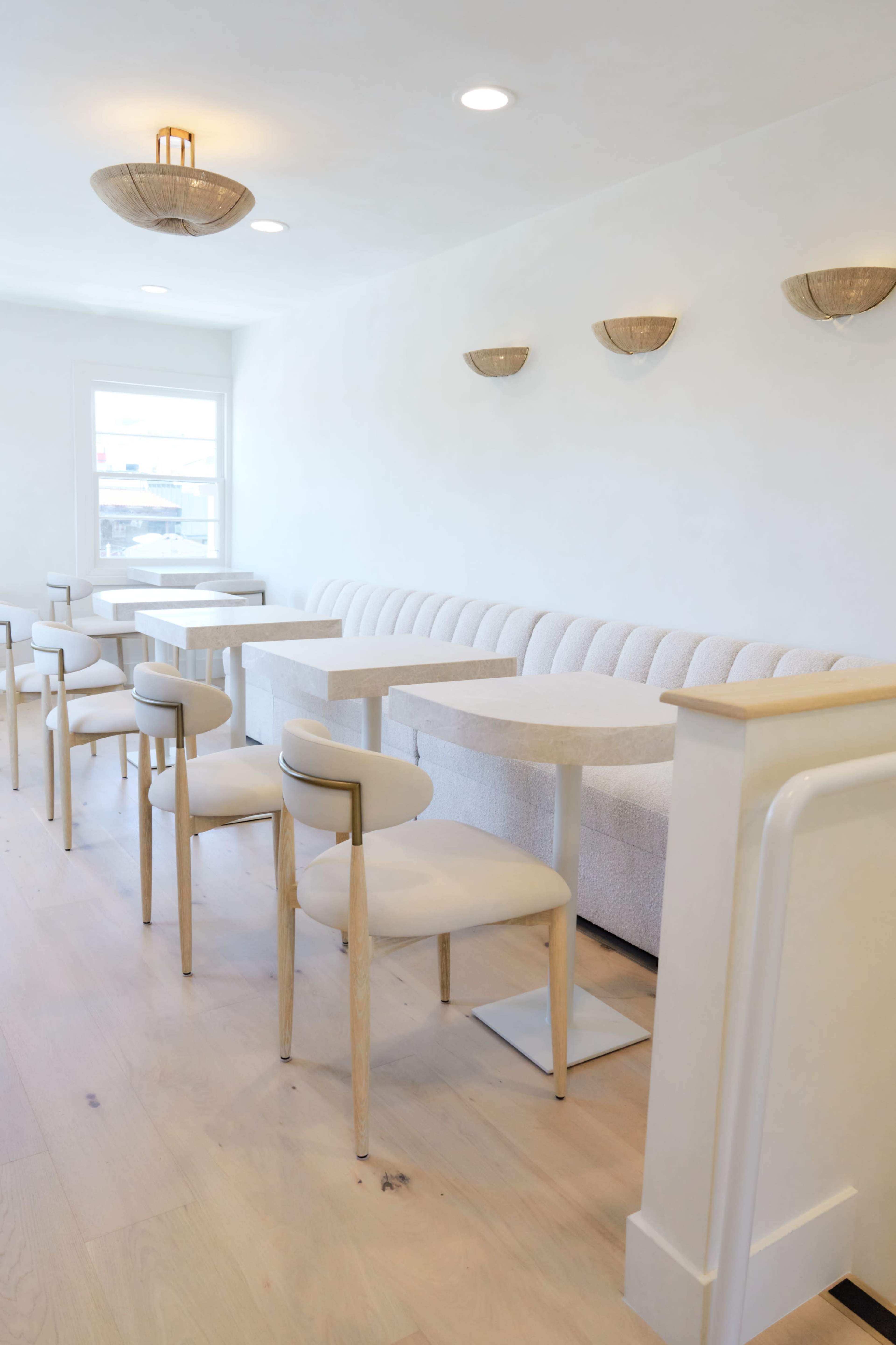 The image shows a bright, modern cafe interior featuring a white sofa, light-colored wooden chairs, and small tables arranged along a light wood floor.