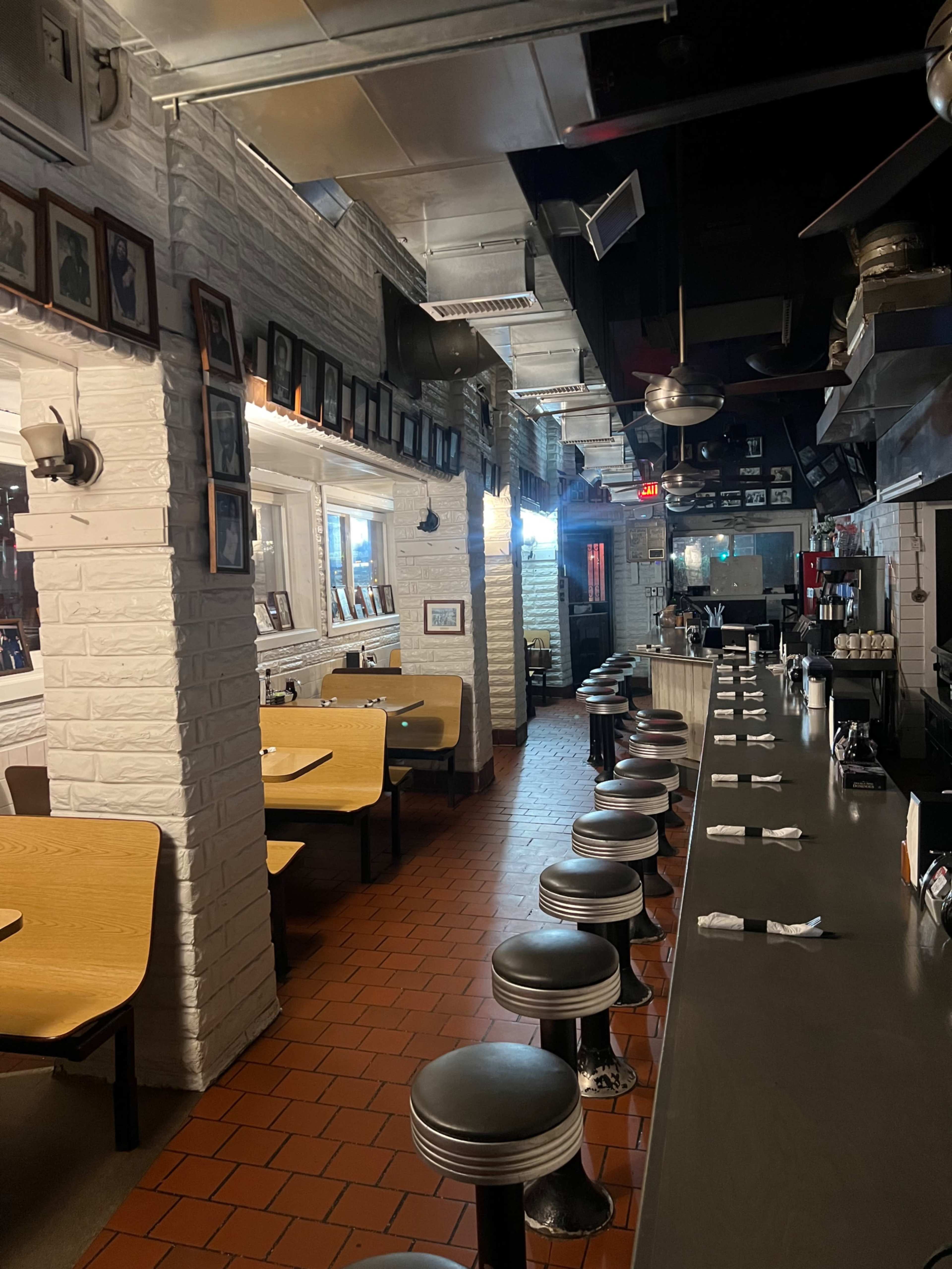 The image shows an empty diner with booths along the walls and a long counter with stools, featuring white brick walls and a tiled floor.
