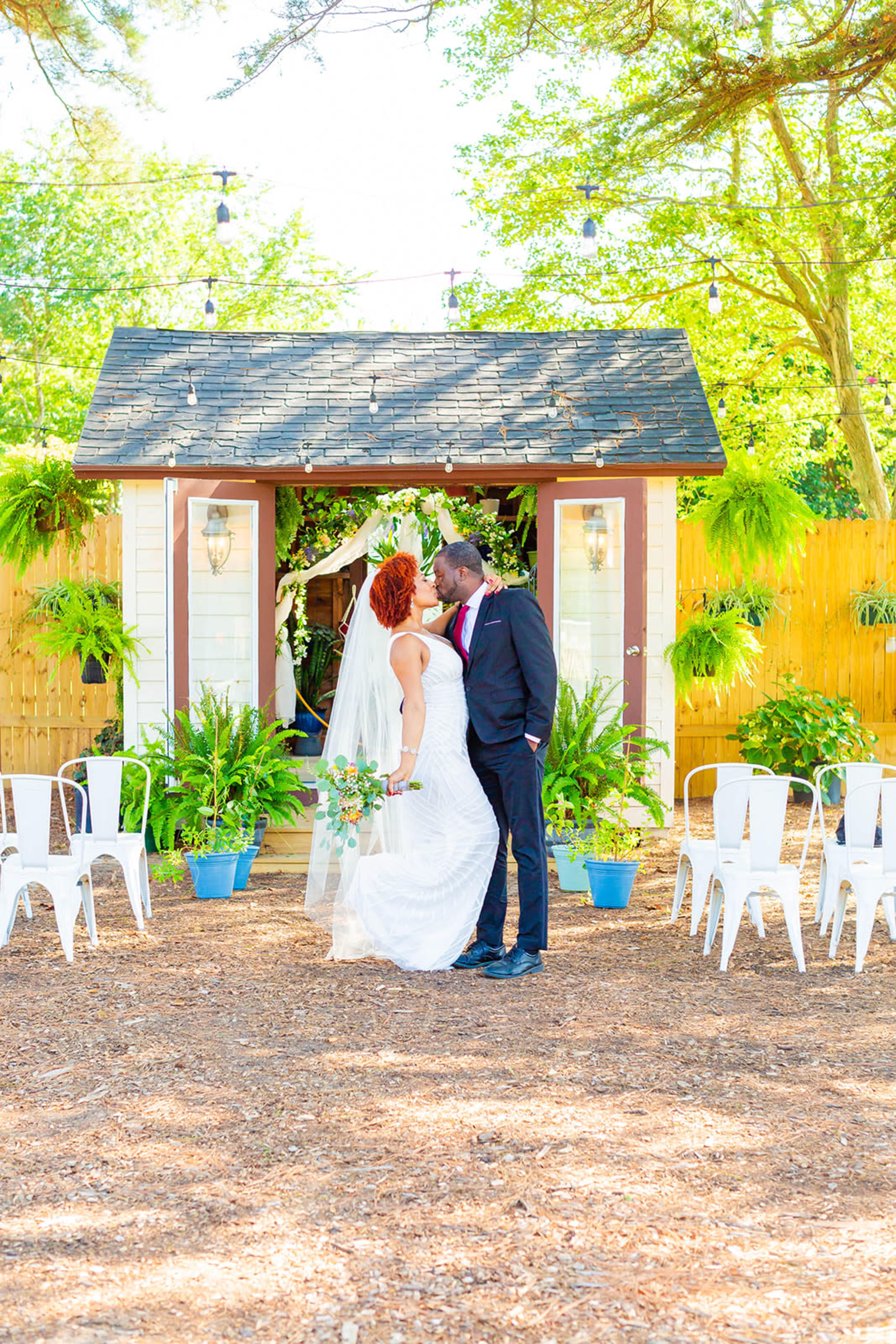 A bride and groom share a kiss under a decorated archway amidst greenery and white chairs in an outdoor wedding setting.