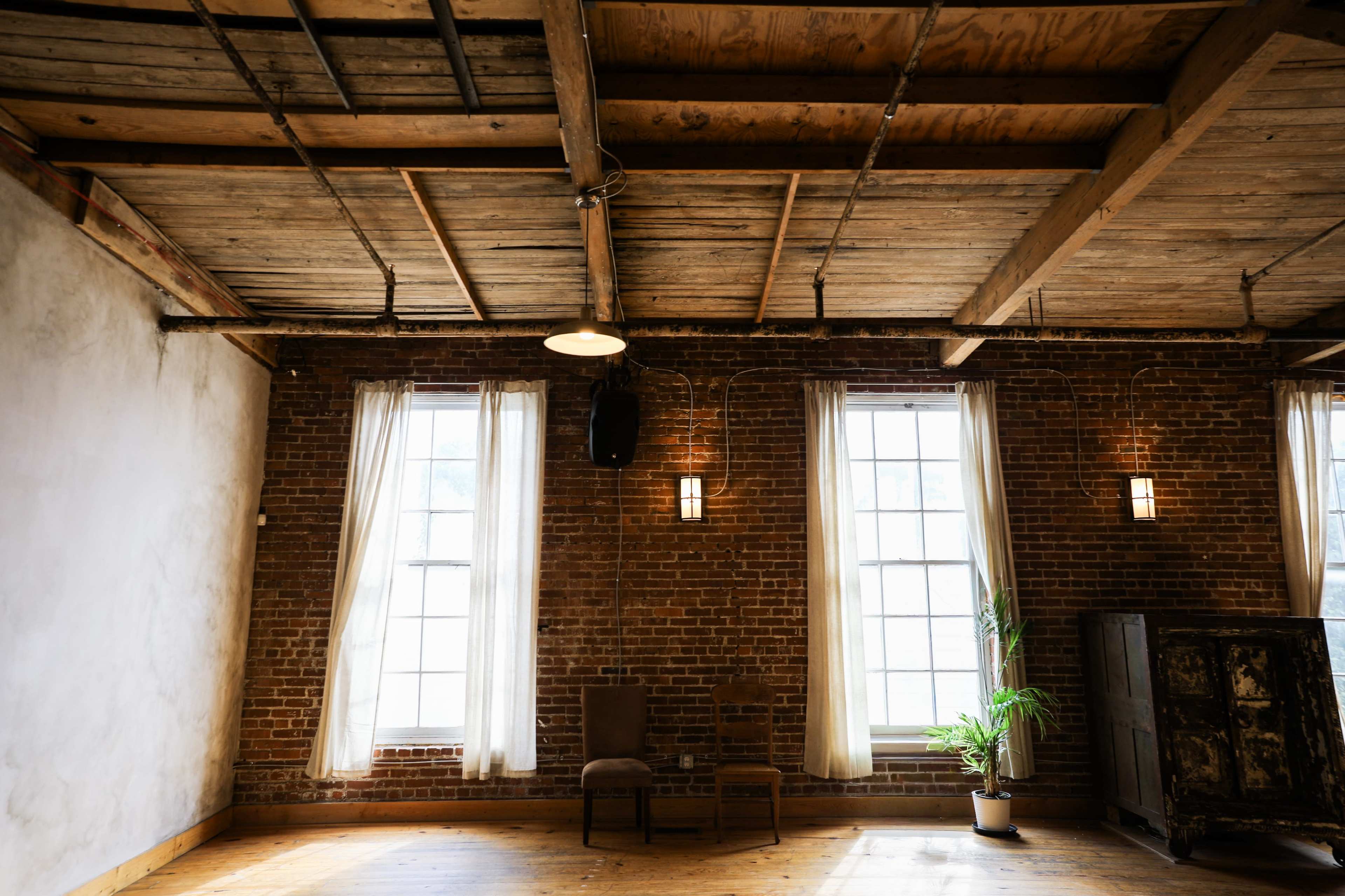 The image shows a spacious room with exposed brick walls, wooden beams, large windows, and minimal furnishings including two chairs and a potted plant.