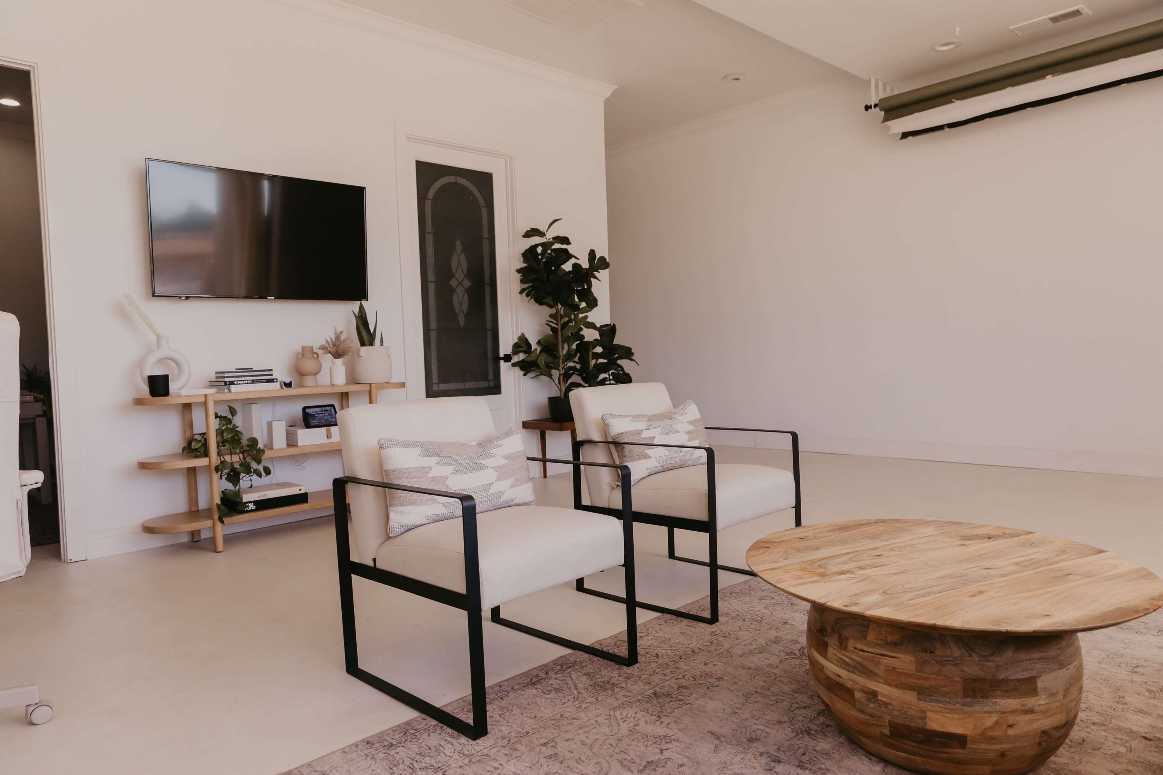 A minimalist living space features two armchairs, a round wooden coffee table, and a wall-mounted television, with plants in the background.