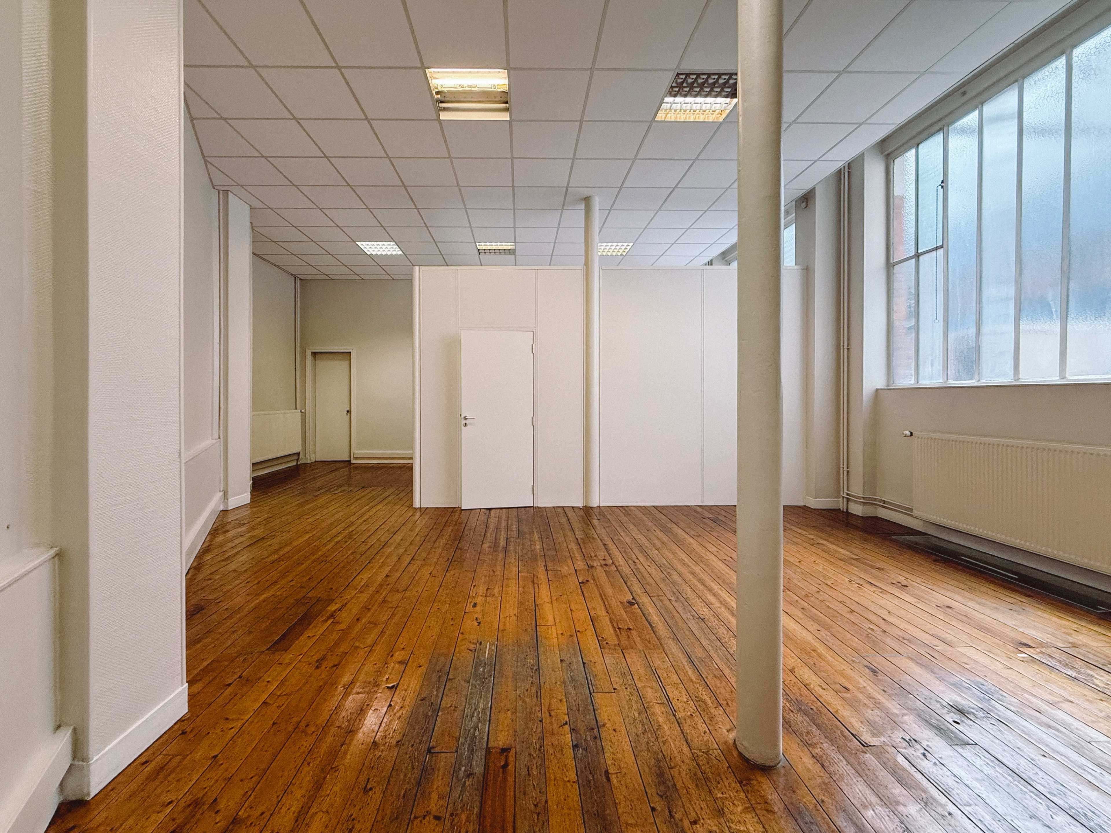 The image shows an empty room with wooden flooring, white walls, and large windows providing natural light.