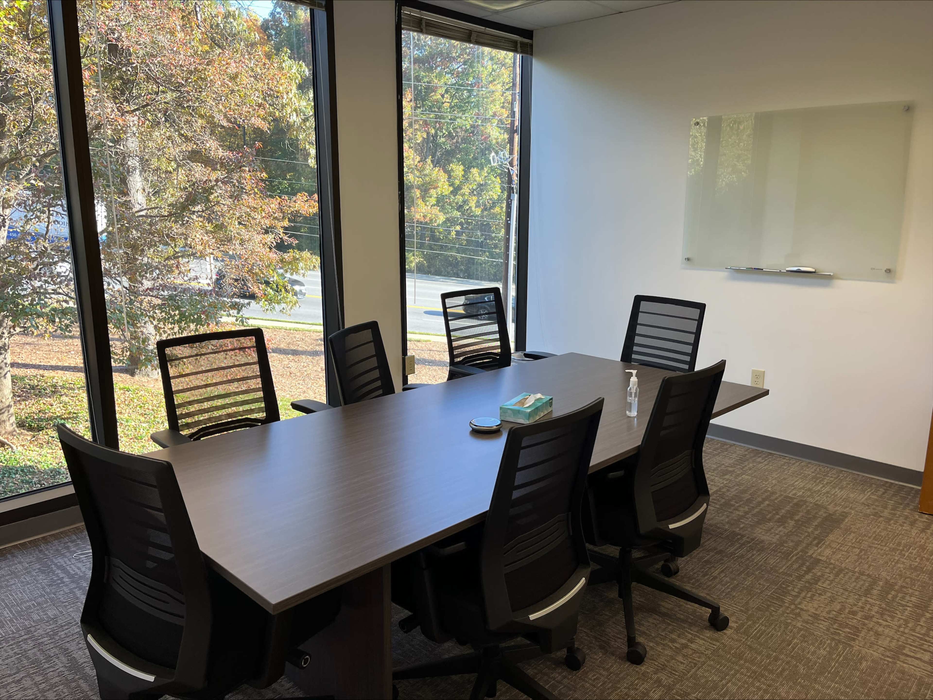 A modern conference room features a large table surrounded by six ergonomic chairs, with large windows showing a view of trees outside.