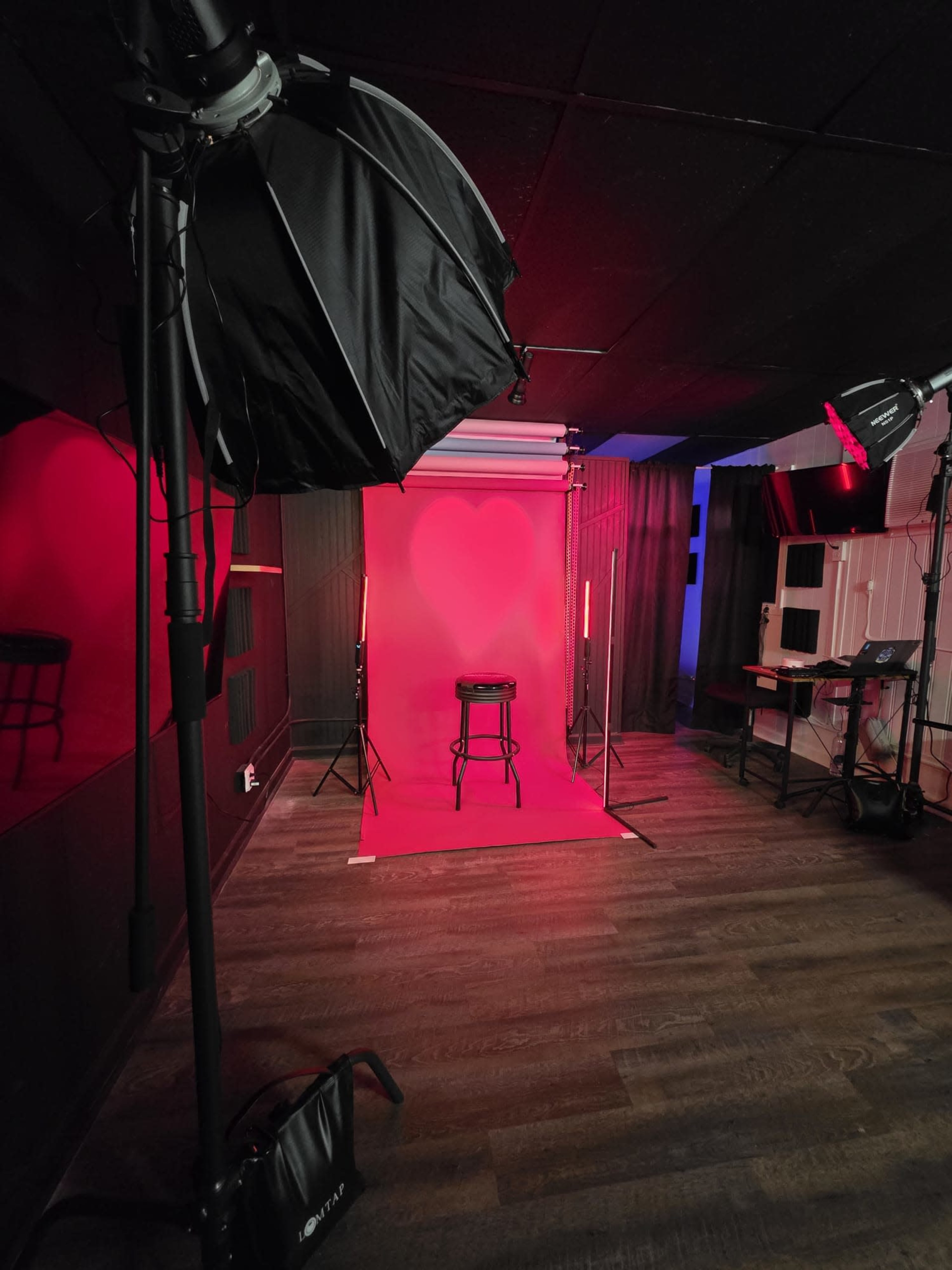 The image shows a photography studio setup with a red backdrop, a black stool in the center, and various lighting equipment positioned around the space.