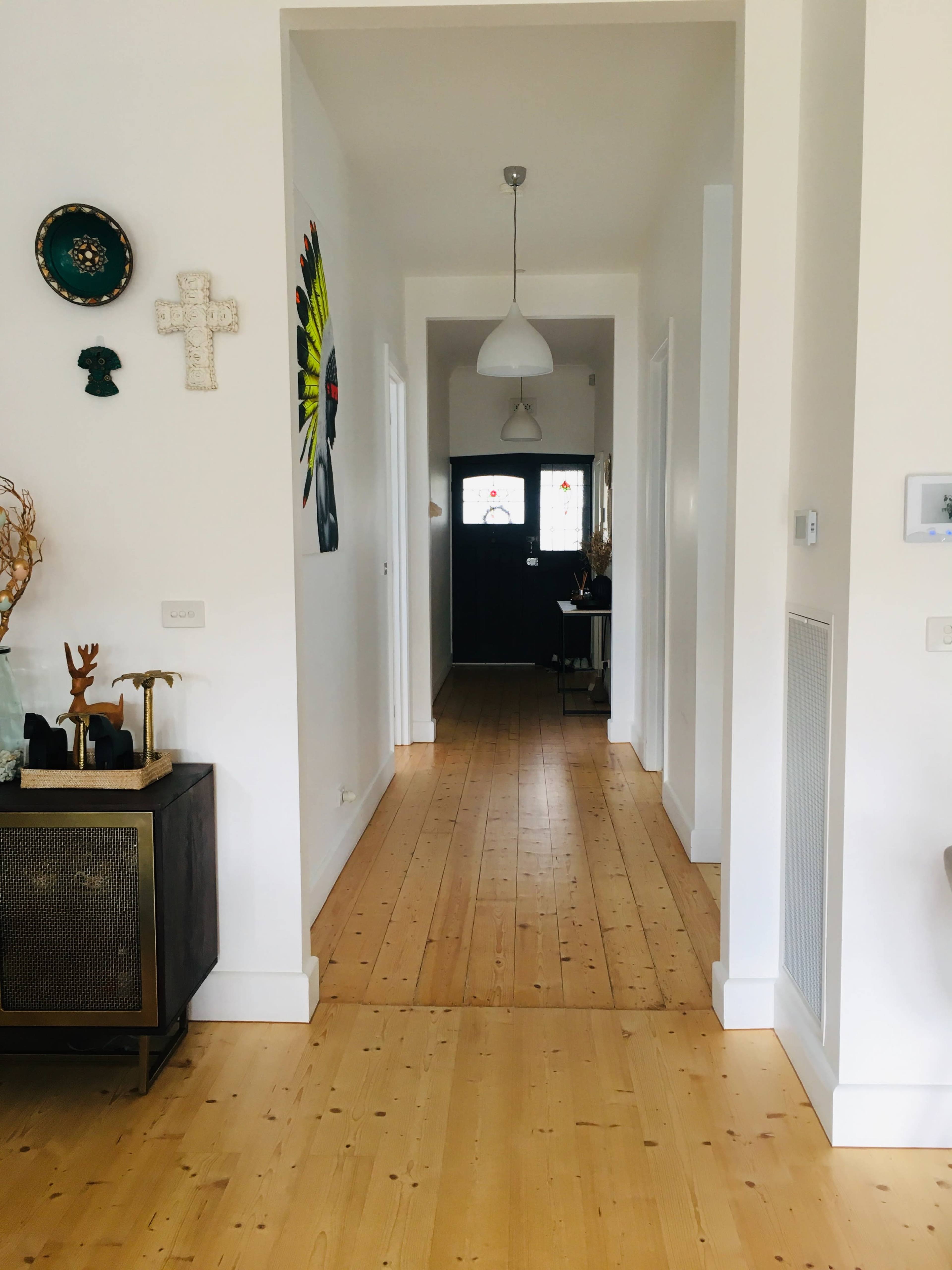 A long, narrow hallway with wooden flooring, framed by white walls and featuring various decorations on one side, leading to a dark door at the end.