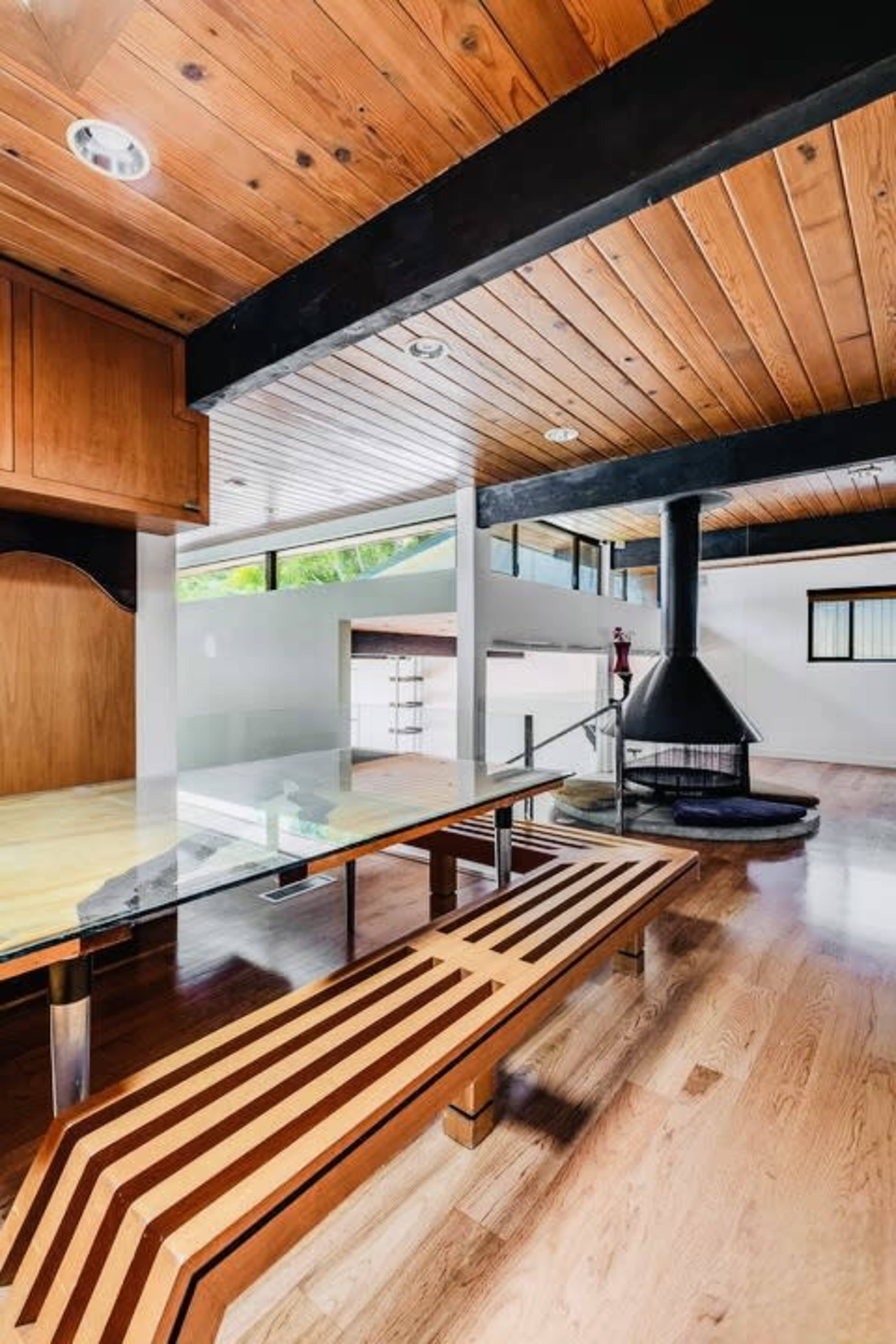 The image shows a modern interior space with wooden ceilings and flooring, featuring a glass-topped table and a black circular fireplace.