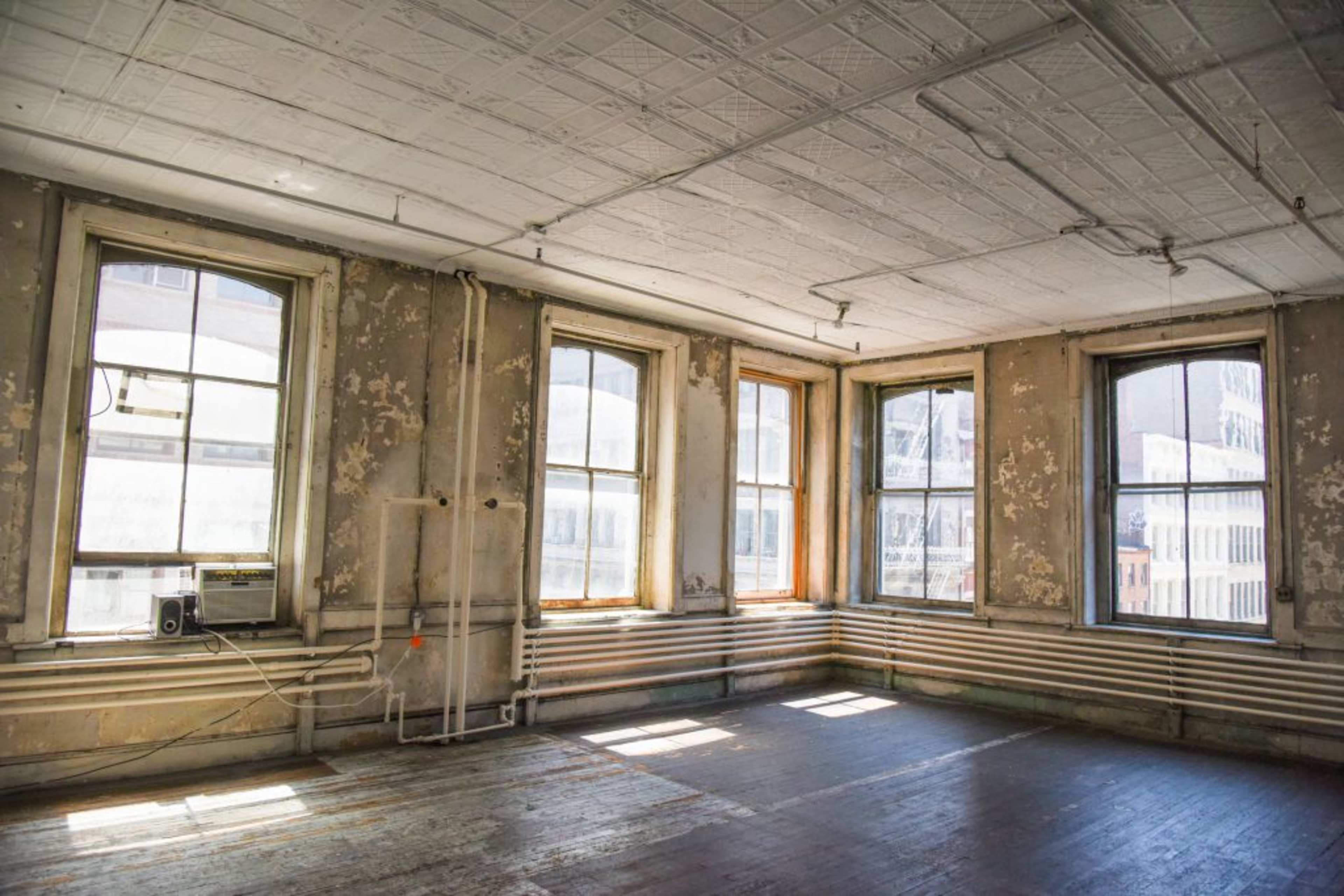 The image shows a sparse, vacant room with three large windows featuring peeling paint and bare wooden flooring.