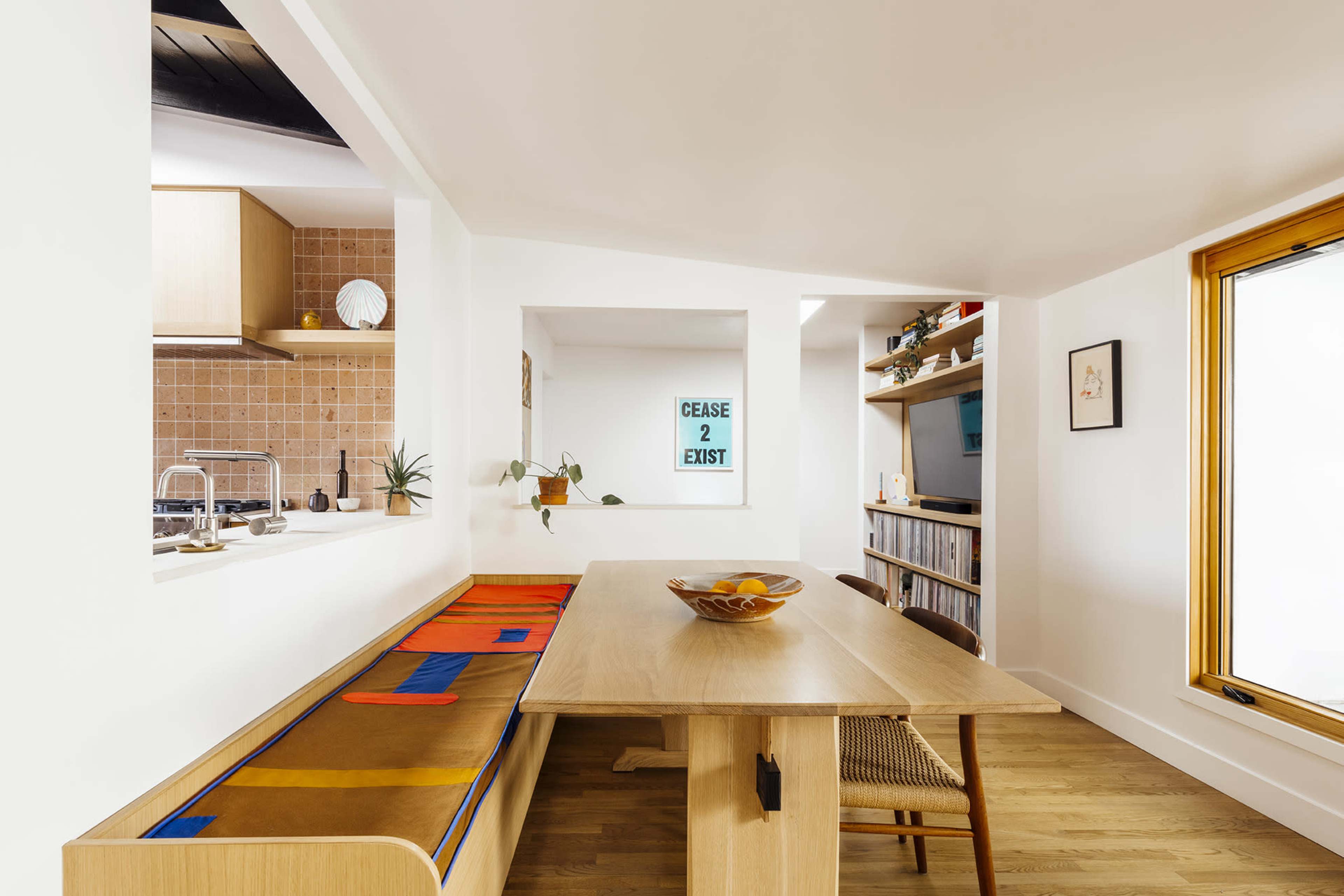 The image shows a modern interior dining area with a wooden table, a bench with colorful cushions, and shelves filled with books, framed by large windows and minimalistic decor.