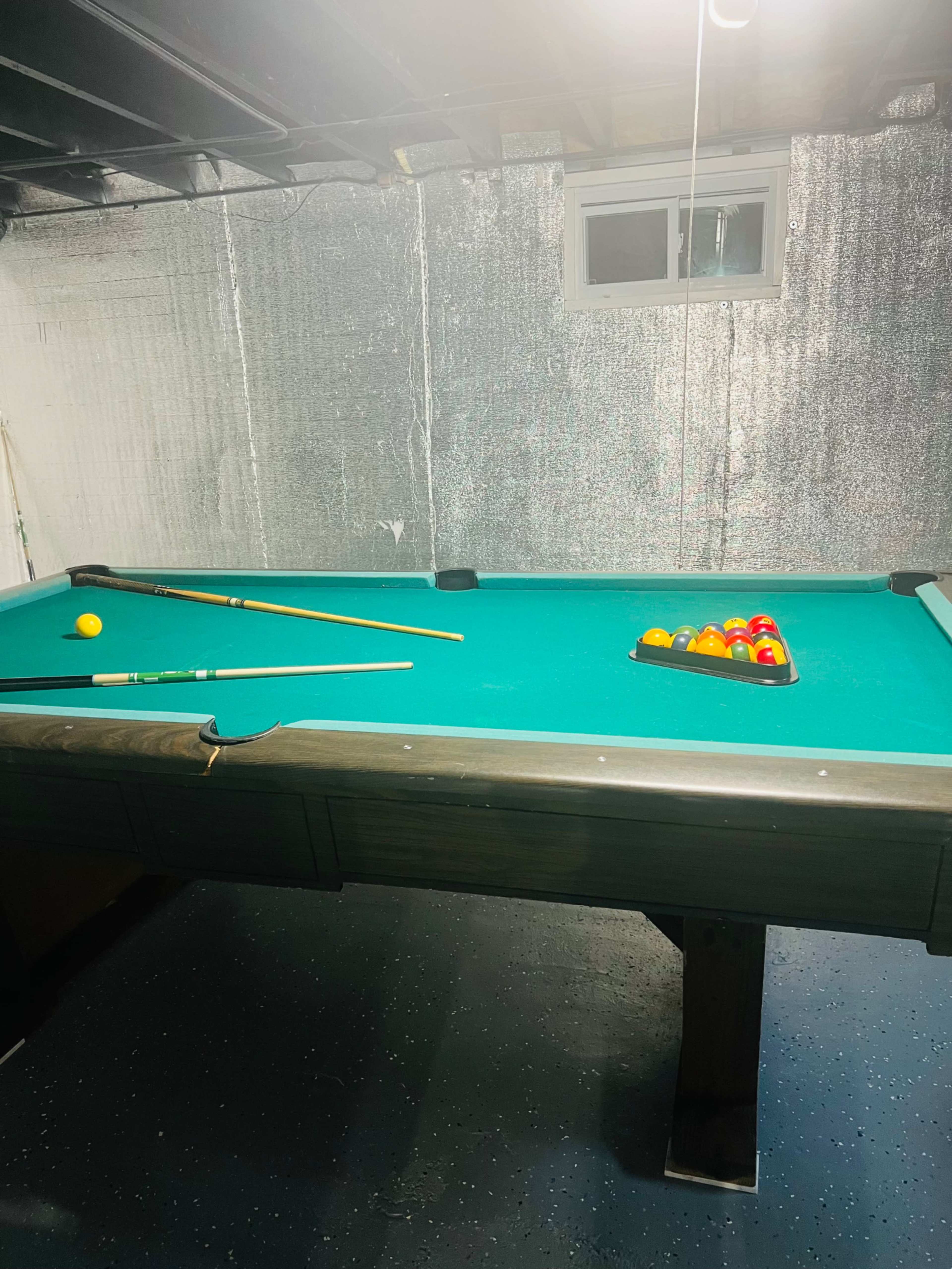 A pool table with a rack of colored balls sits in a dimly lit basement.