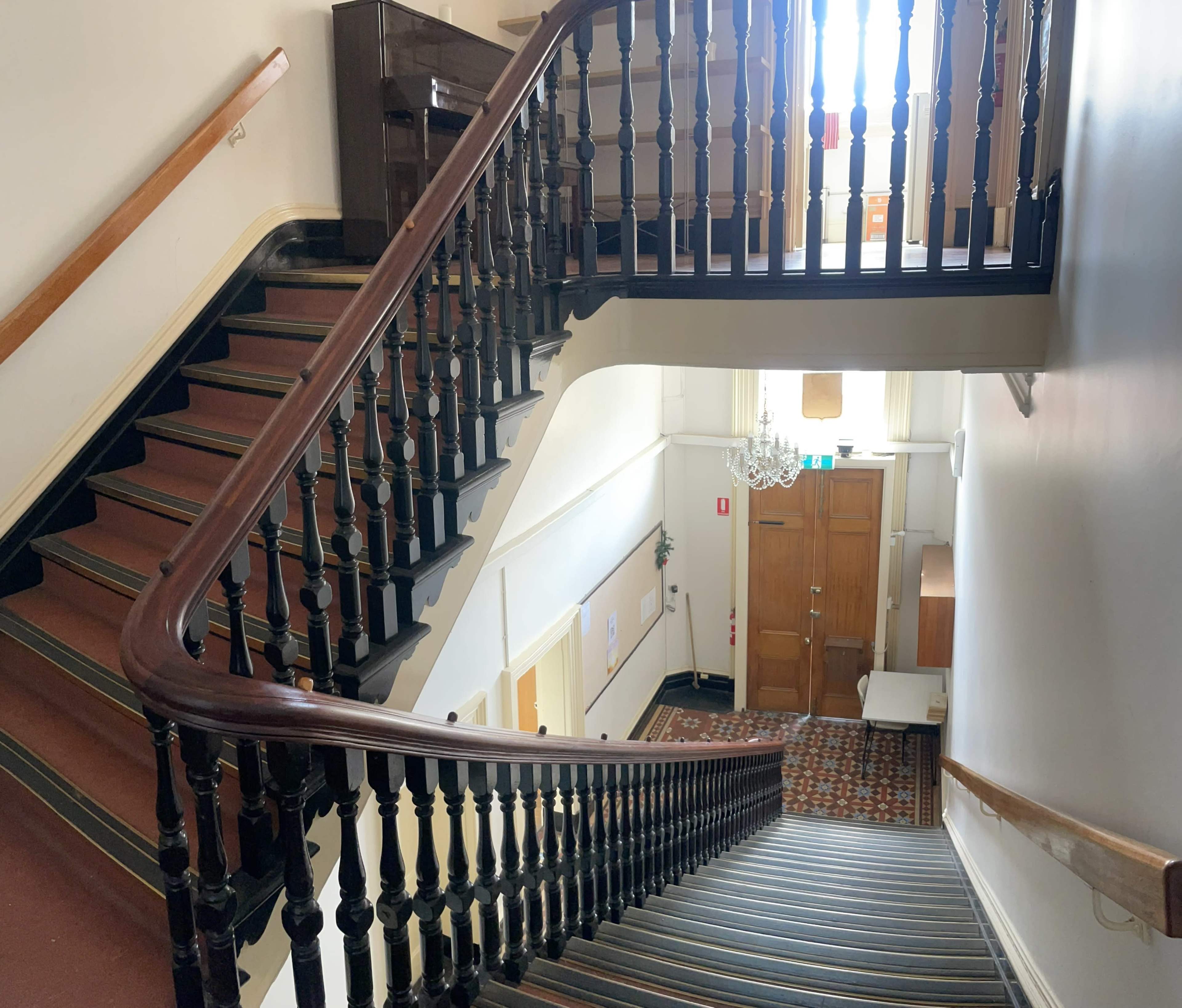 A staircase with a wooden railing leading down to a landing, with a chandelier hanging above and a wooden door at the bottom.