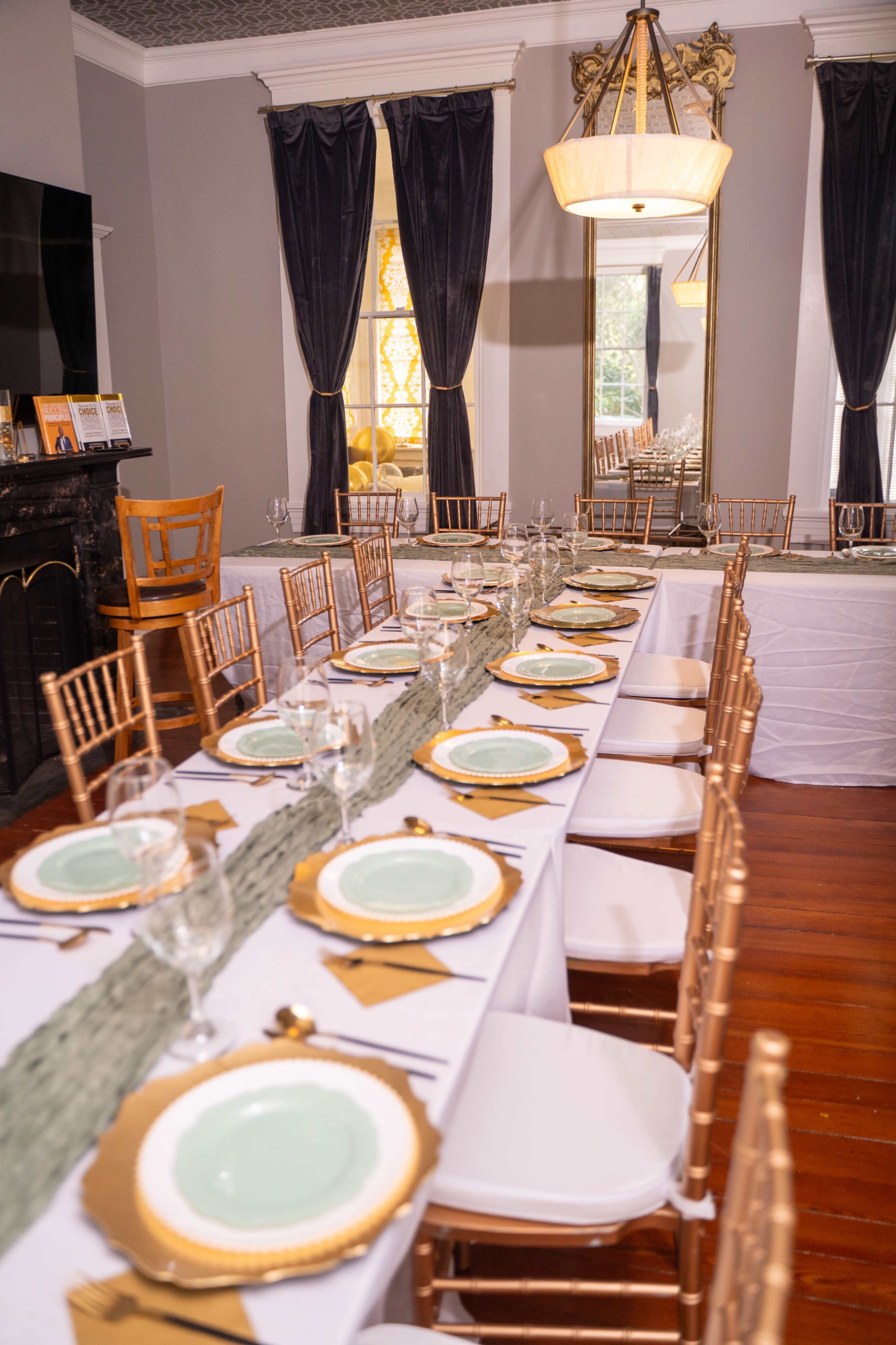 A long dining table is elegantly set with mint green plates and gold accents, surrounded by gold chiavari chairs in a room with gray walls and dark curtains.