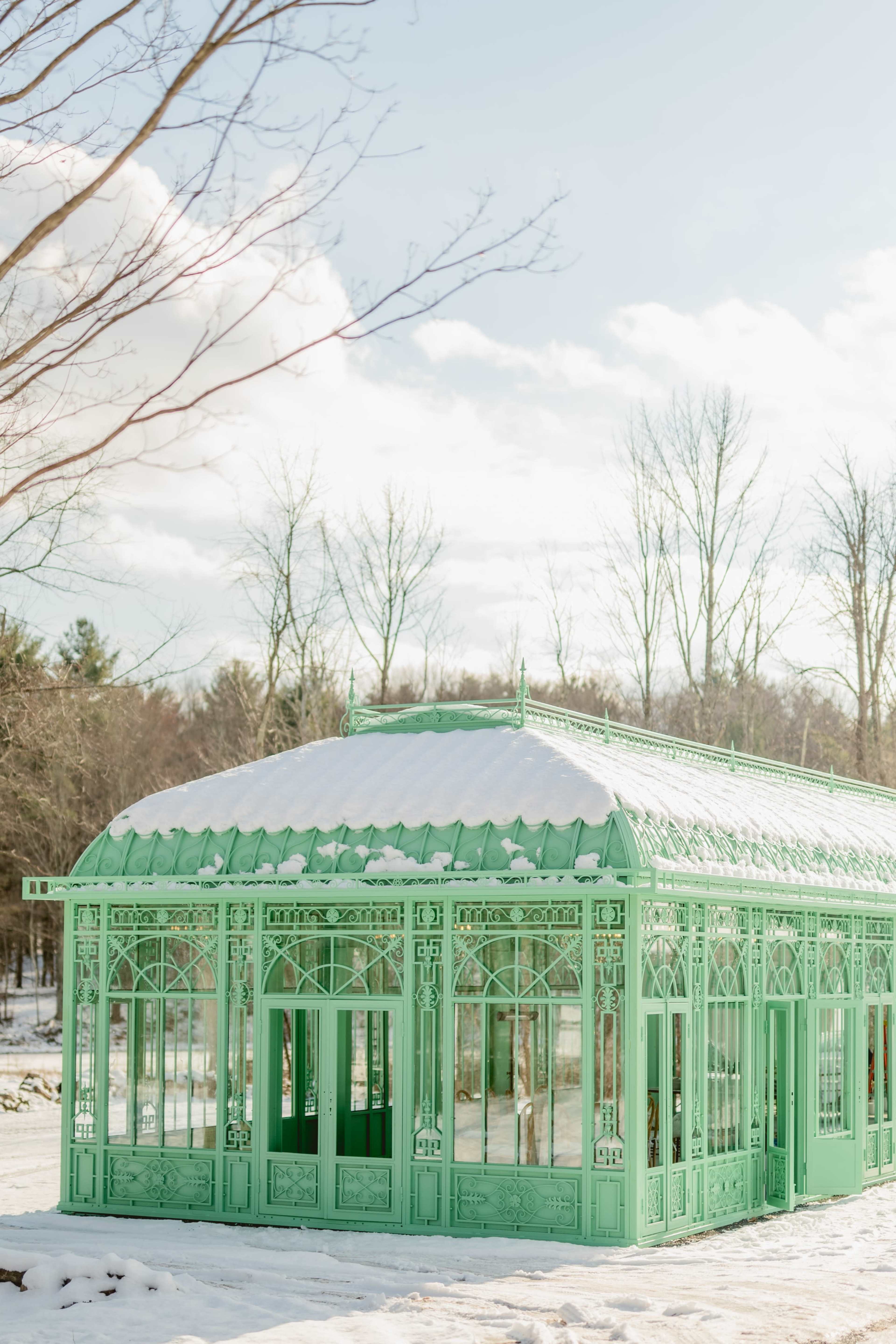 A green glass greenhouse sits on a snowy landscape, surrounded by bare trees.