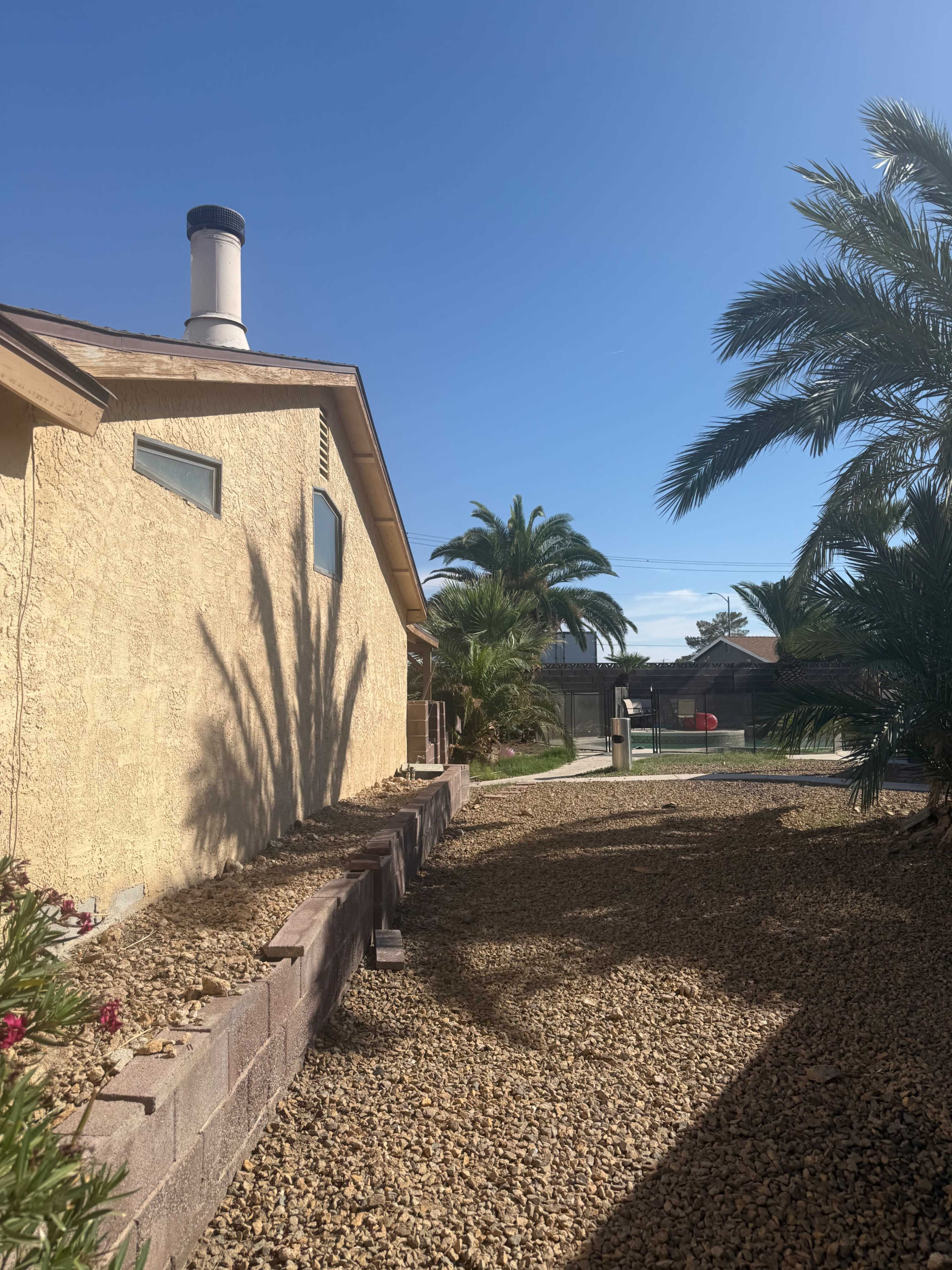 A gravel path runs alongside a house, bordered by a palm tree and a wall, leading to a backyard with a sports court.