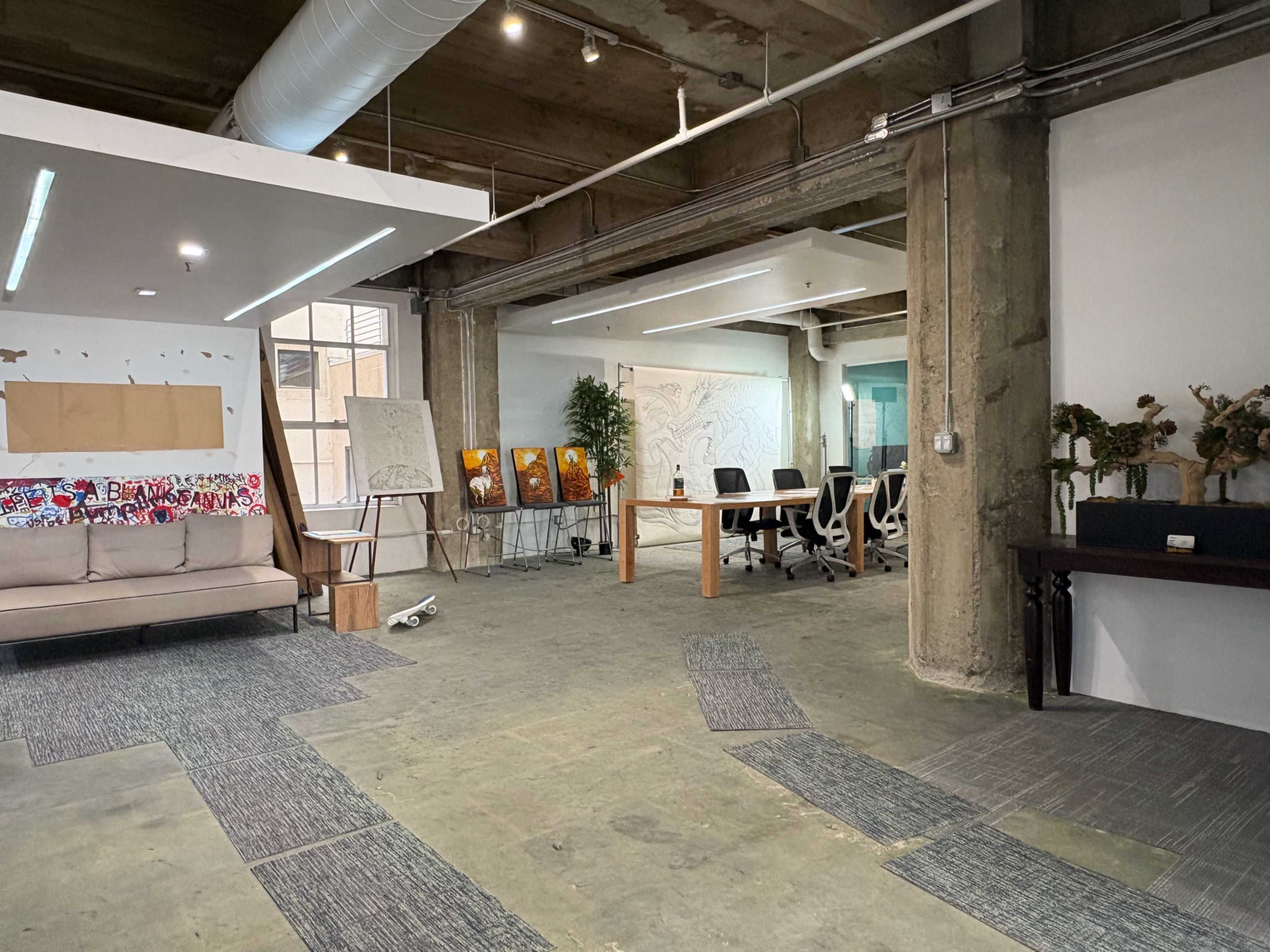 The image shows a spacious office area with a sofa, a large table surrounded by chairs, and artworks displayed on easels against a concrete wall.