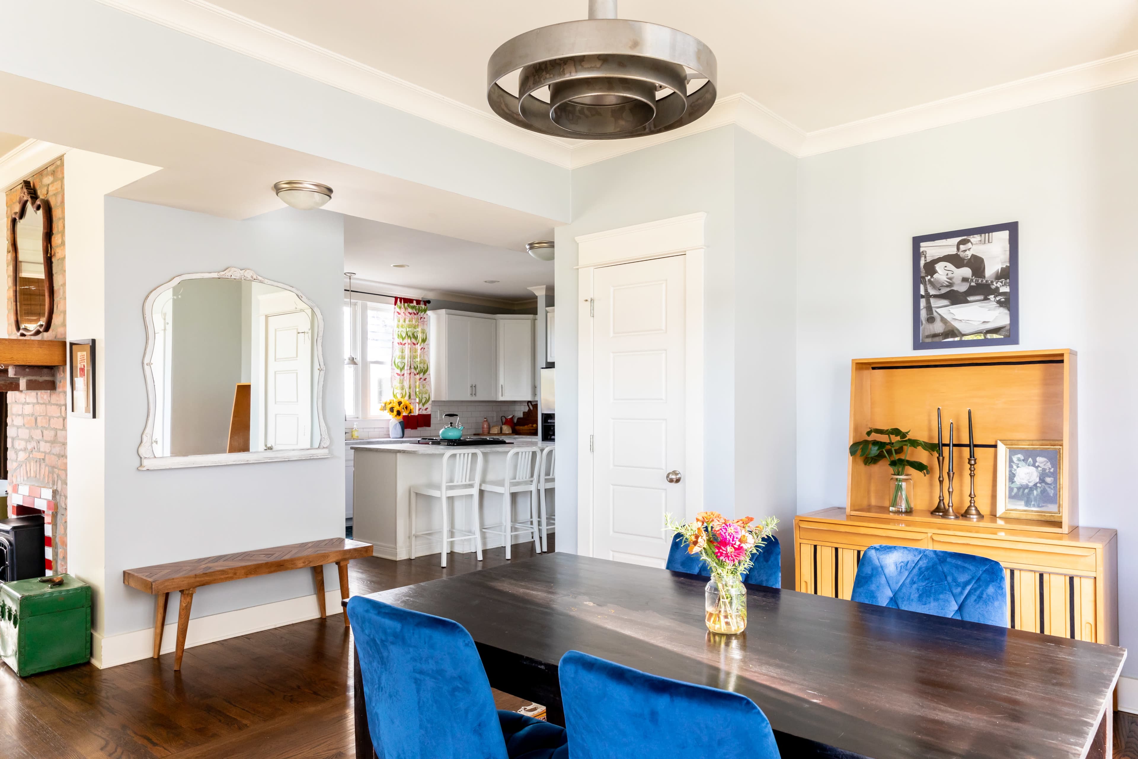 The image shows a dining area with a large dark table and blue chairs, leading into a kitchen with white cabinetry and a mirror on the wall.