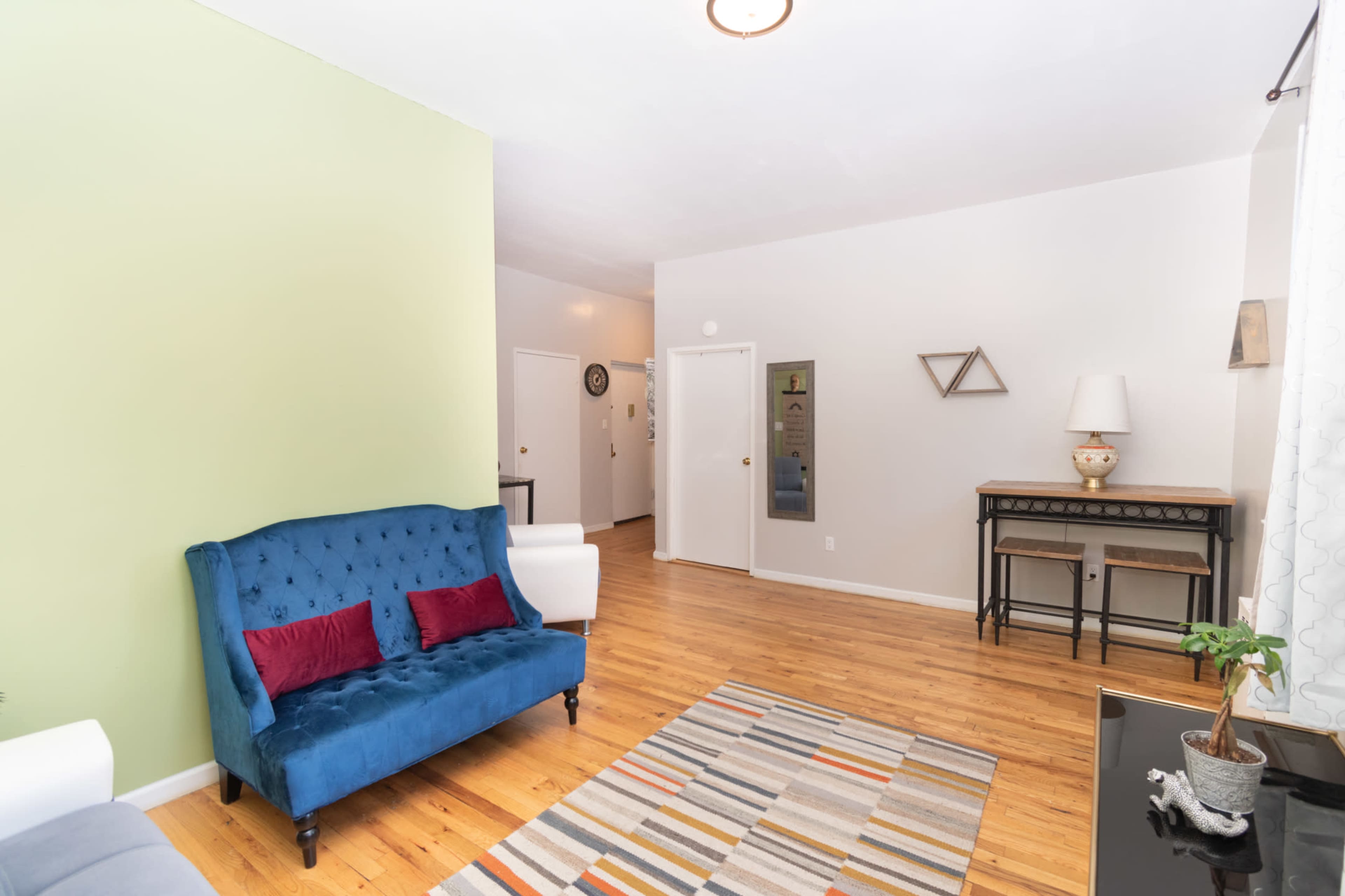 The room features a blue velvet couch, a striped rug, wooden flooring, and a small table with a lamp against a light-colored wall.