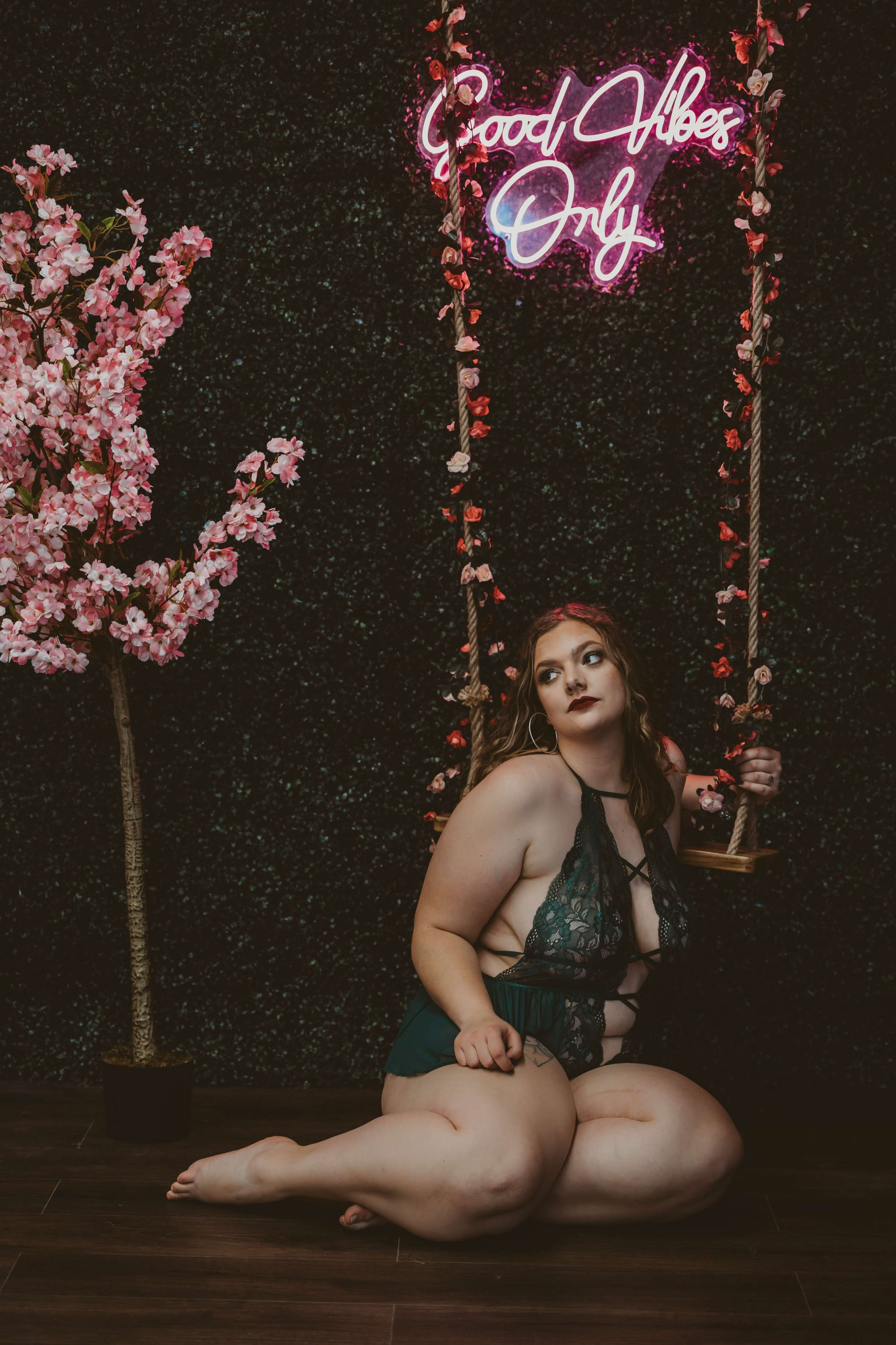 A woman in a green lace outfit poses on a swing near a wall decorated with artificial flowers and a neon sign that reads "Good Vibes Only."