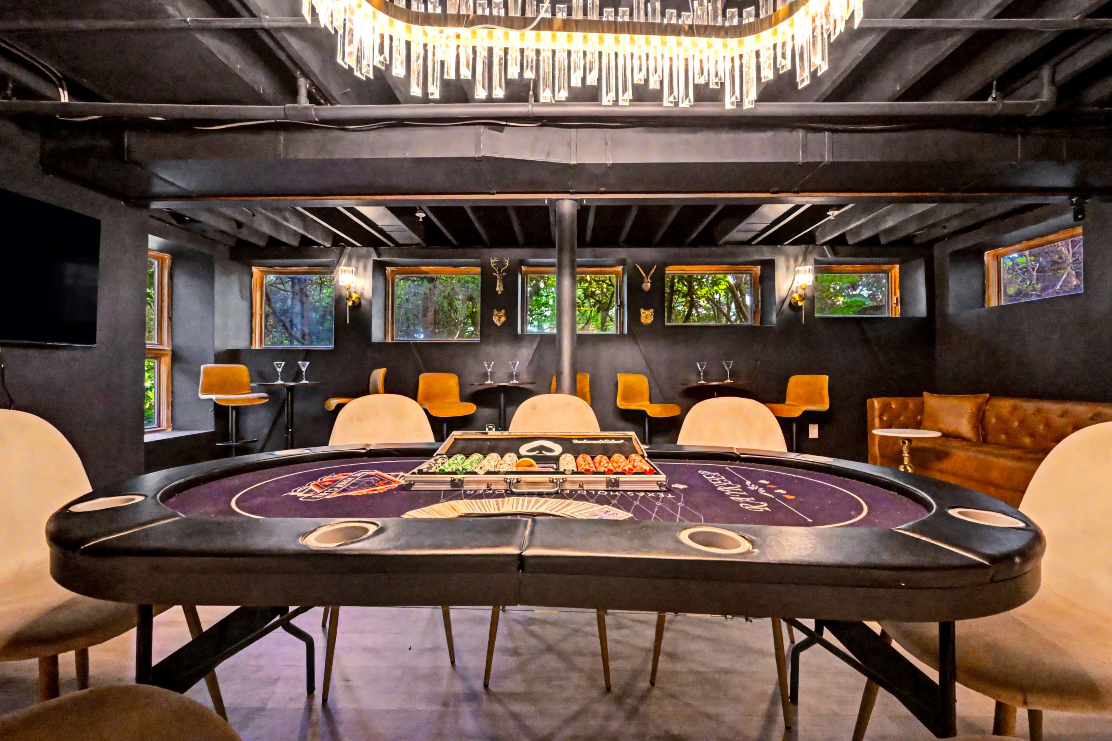 The image shows a stylish poker room with a large round table, a poker set in the center, and modern seating arrangements surrounded by large windows.